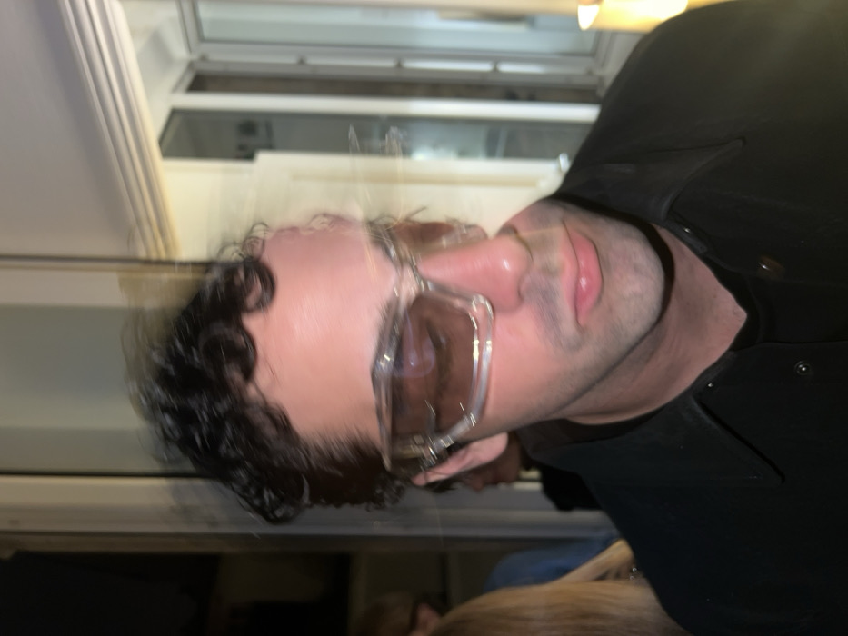 A male individual, appearing to be in his 30s-40s, with dark, curly hair and light stubble, is depicted in a close-up, slightly off-center. He wears clear-framed sunglasses with brown-tinted lenses. His attire consists of a dark, collared shirt or jacket over a dark undergarment. The individual's face and head exhibit significant motion blur, indicating movement during exposure. He maintains a neutral expression with slightly parted lips, gazing slightly downward and to his left. The scene is set indoors, characterized by dim lighting suggestive of an evening event within an indoor venue in Paris, France. Architectural elements include a tall, light-framed window or door structure visible in the blurred background to the right, reflecting interior light. A warm, artificial light source emits a glow on the extreme right. Partially visible in the blurred background to the left are other individuals, including a person with blonde hair wearing a blue top.