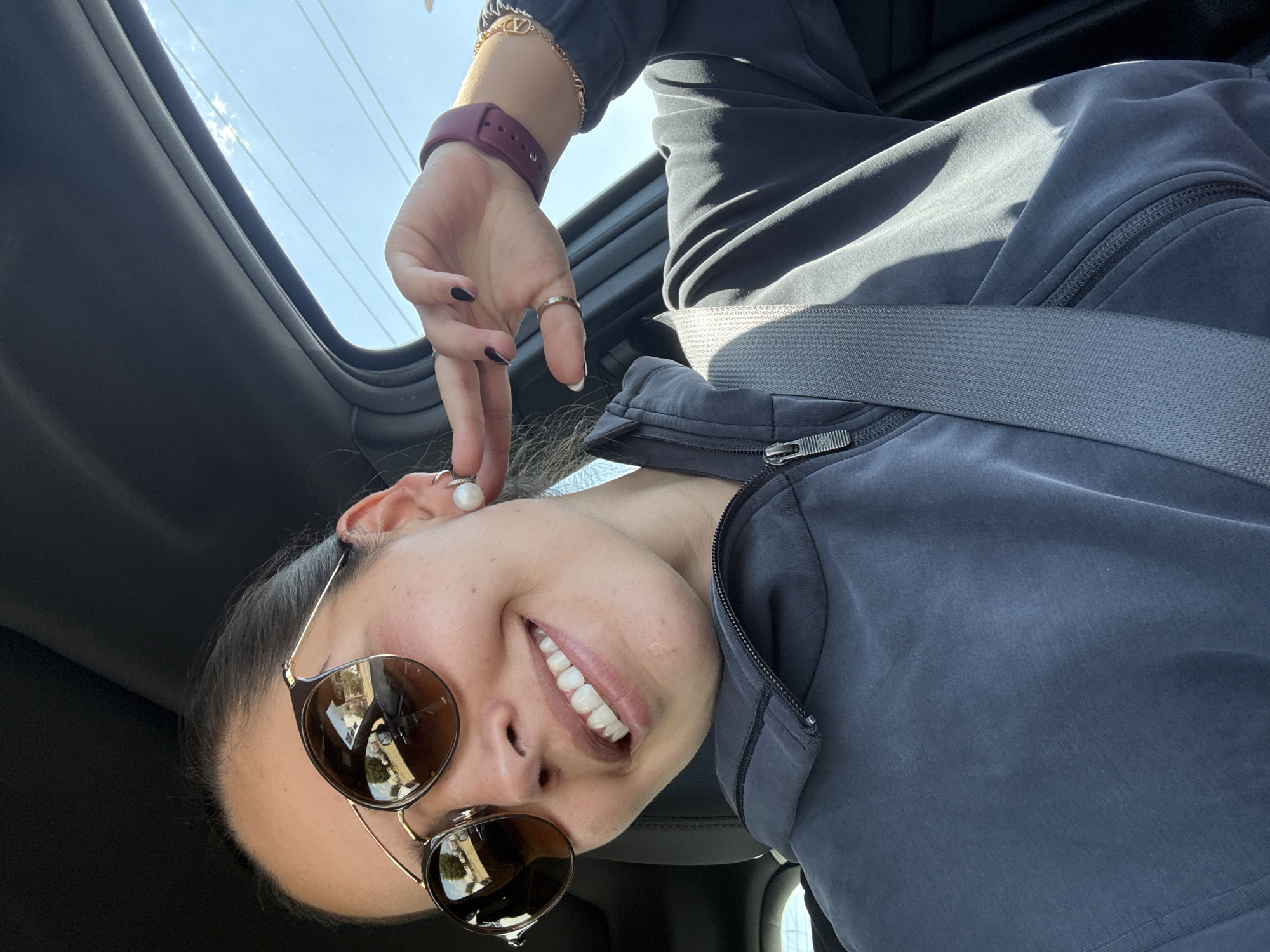 A female is depicted in the driver's seat of a vehicle on a clear, sunny day in San Bernardino Tlaxcalancingo, Mexico. She is observed from the chest up, wearing dark aviator-style sunglasses that reflect trees and a bright sky. Her hair is pulled back. She displays a broad smile, revealing white teeth. She is dressed in a dark gray hooded sweatshirt with a visible zipper and is secured by a gray shoulder seatbelt. Her left hand is raised, with fingers touching a large white pearl stud earring on her right ear. On her left wrist, she wears a maroon-colored smartwatch and a gold-colored chain bracelet with a distinct 'V' charm. A silver-colored ring is present on her left thumb, and her fingernails on the left hand are painted a dark shade. The vehicle's interior appears dark-toned. Through the upper right window, a clear blue sky with sparse clouds and horizontal utility lines is visible.