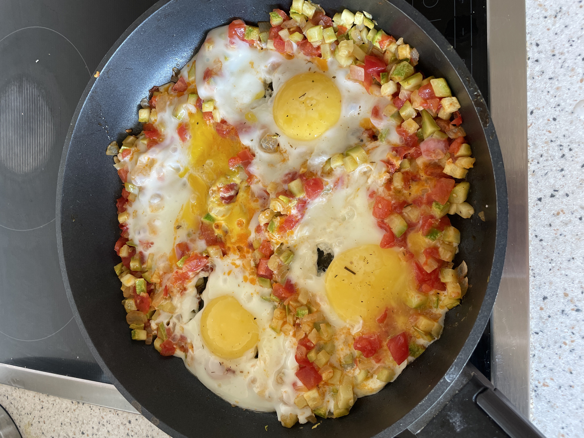 A top-down shot shows a frying pan containing eggs cooked with diced vegetables. The pan is black and sits on a black stovetop with a light speckled countertop surrounding it. The eggs are mostly cooked, with some yolks intact and some broken. The diced vegetables appear to be primarily zucchini and tomato. The scene appears to be indoors. The location is Бордуки, Russia.