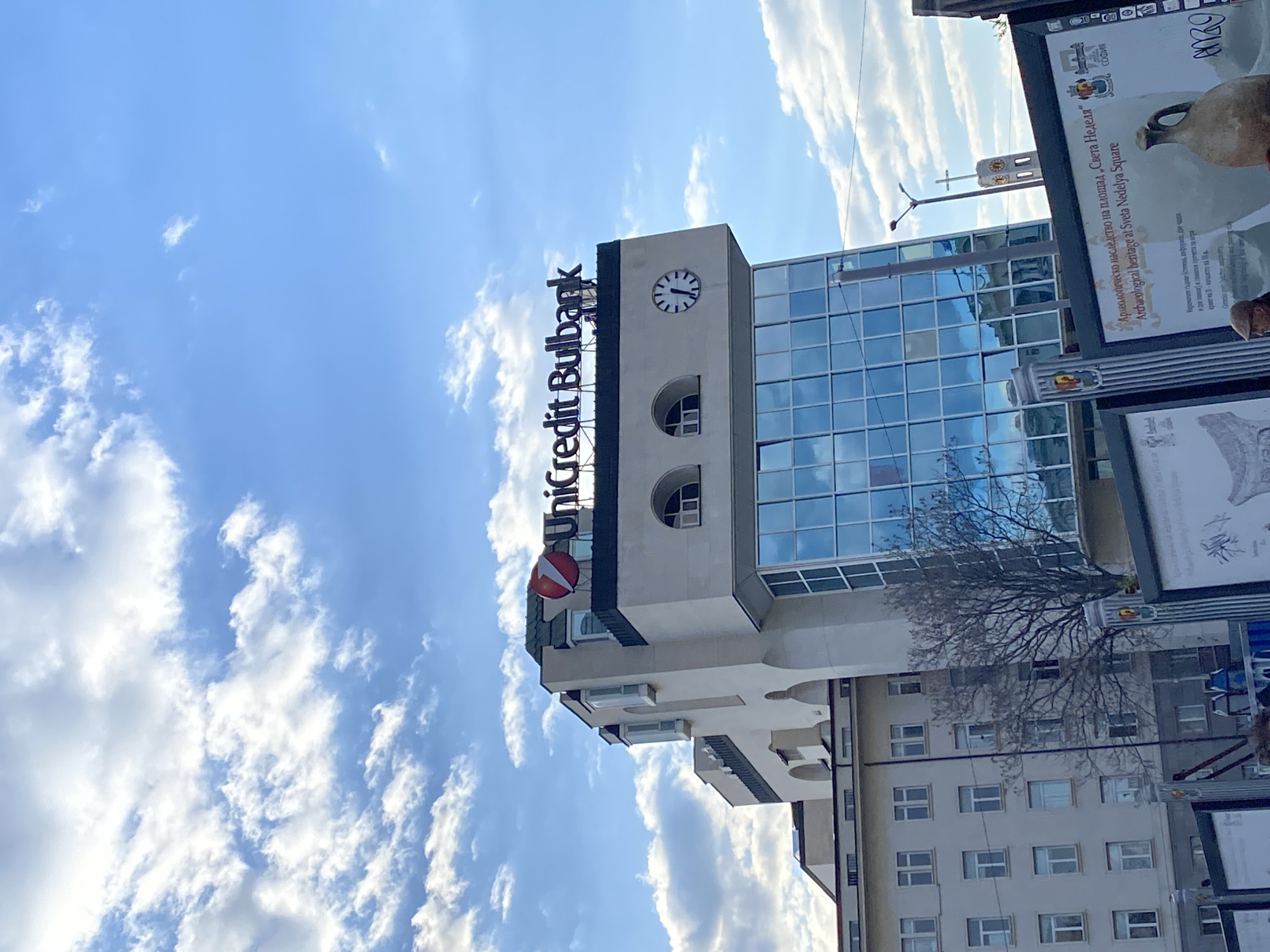 The image captures a vibrant cityscape, likely in Sofia, Bulgaria, dominated by the UniCredit Bulbank building under a bright blue sky dotted with scattered white clouds. The building features the prominent "UniCredit Bulbank" sign at its top. Below the signage, a round clock face displays the time as approximately 7:15. The upper floors of the building boast a modern glass facade that reflects the sky, while lower sections are made of lighter-colored material with both arched and rectangular windows.
To the right of the UniCredit building, large informational panels are visible. Text on one of these panels, in both Bulgarian and English, reads: "Археологическо наследство на площад „Света Неделя" Archaeological heritage at Sveta Nedelya Square," indicating the specific location. The city "СОФИЯ" is also visible, along with the number "2000." A bare-branched tree stands in front of another building with multiple windows, contributing to the urban setting. The overall lighting suggests it is daytime, possibly morning given the clear sky and the time displayed on the clock. No people are visible in the frame.
