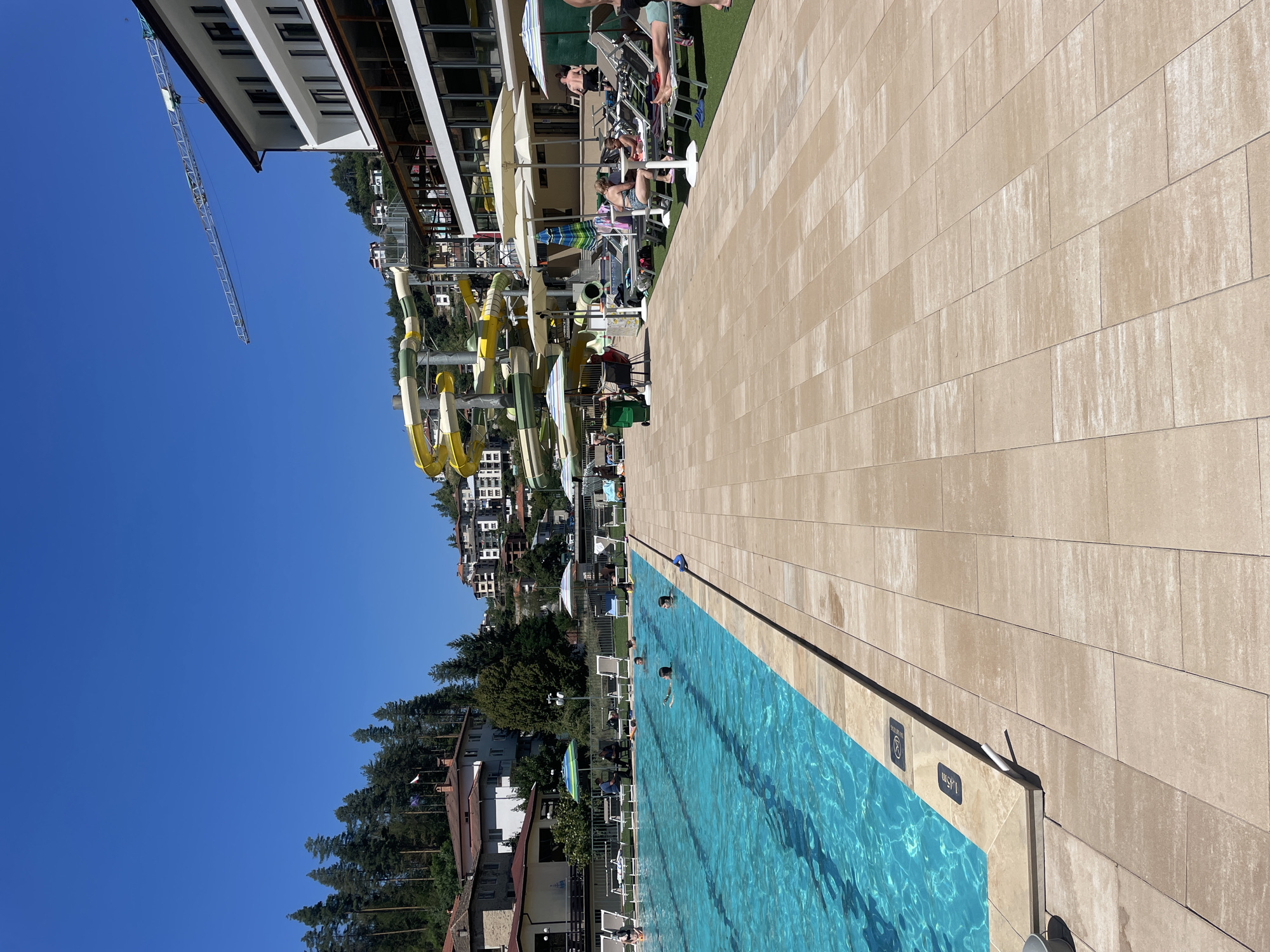 This image depicts a resort area in Banite, Bulgaria. In the foreground, a large, rectangular swimming pool with clear blue water is visible. Several people are swimming or wading in the pool. The pool area is paved with light-colored square tiles, and several lounge chairs are arranged on the tiles. In the background, there are several buildings, including a hotel with a white facade and balconies. A colorful water slide structure is also present, with yellow and green sections. Beyond the buildings, a densely forested hillside rises, dotted with more residential buildings. The sky is a clear, bright blue, and a construction crane is visible in the upper right portion of the image, suggesting ongoing development. Umbrellas are set up around the pool area, providing shade for sunbathers.