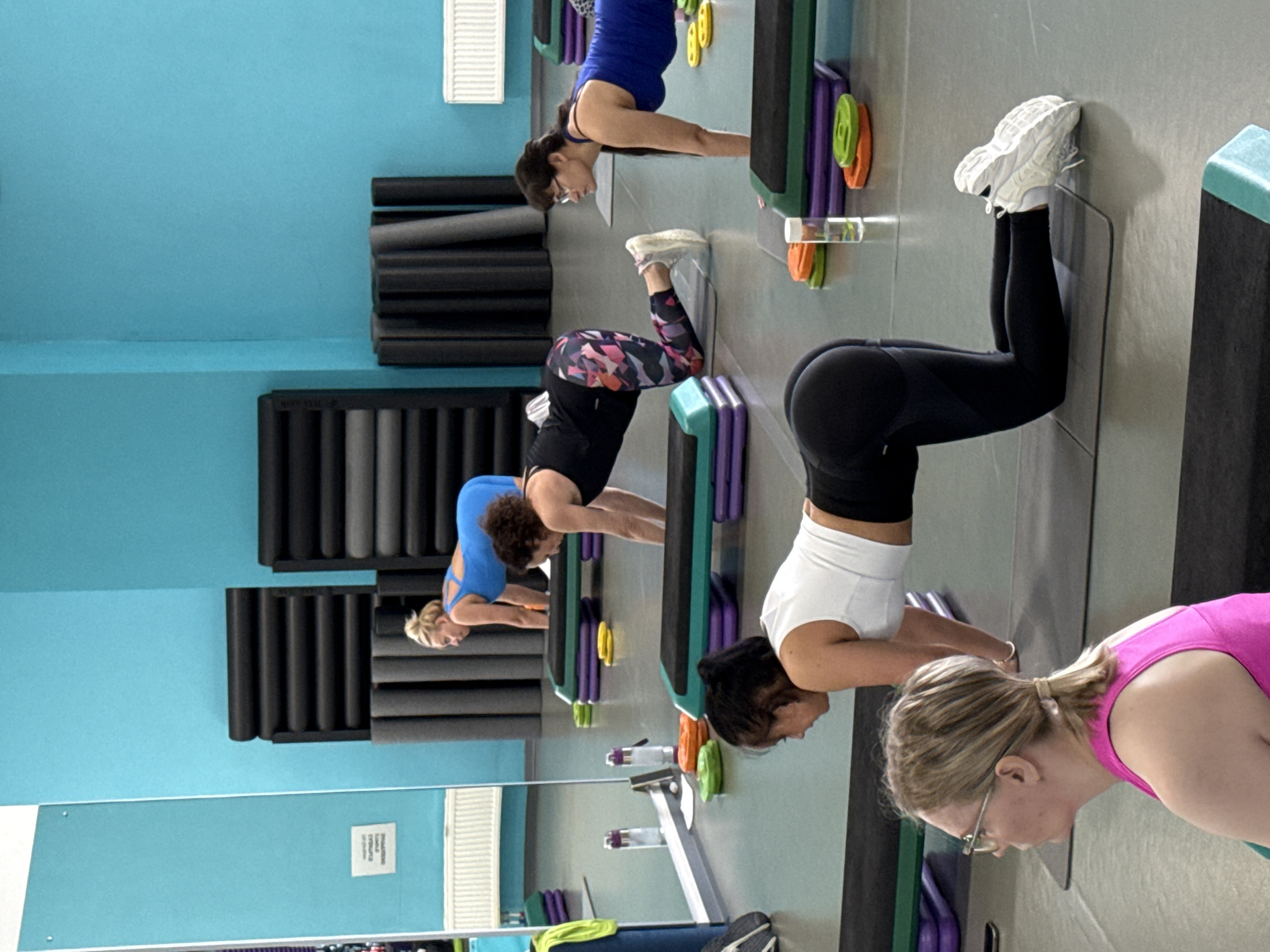 The image captures a group of women engaged in a fitness class within a gym or studio in Yekaterinburg, Russia. The setting features light blue walls, a light gray floor, and mirrored surfaces reflecting parts of the room.
Several women are visible, all dressed in athletic wear including leggings, sports tops, and sneakers. Their postures suggest they are performing a synchronized exercise routine, possibly a form of stretching, warm-up, or a step aerobics drill. Most women are bent forward at the waist, with their hands placed on step platforms or the floor. Their hair is generally tied back, and some wear glasses, indicating focus on their activity.
Prominent objects in the scene include numerous step aerobics platforms, visible in green, purple, and black with colorful risers (yellow, orange, green). Along the back wall, there are racks storing black and gray foam rollers or yoga mats. Water bottles are also seen near some of the exercise stations. The overall atmosphere appears to be one of concentrated physical activity.
On a lower section of the wall, to the left, a small white rectangular sign with dark, illegible text is visible. The lighting is bright and even, typical of an indoor gym.