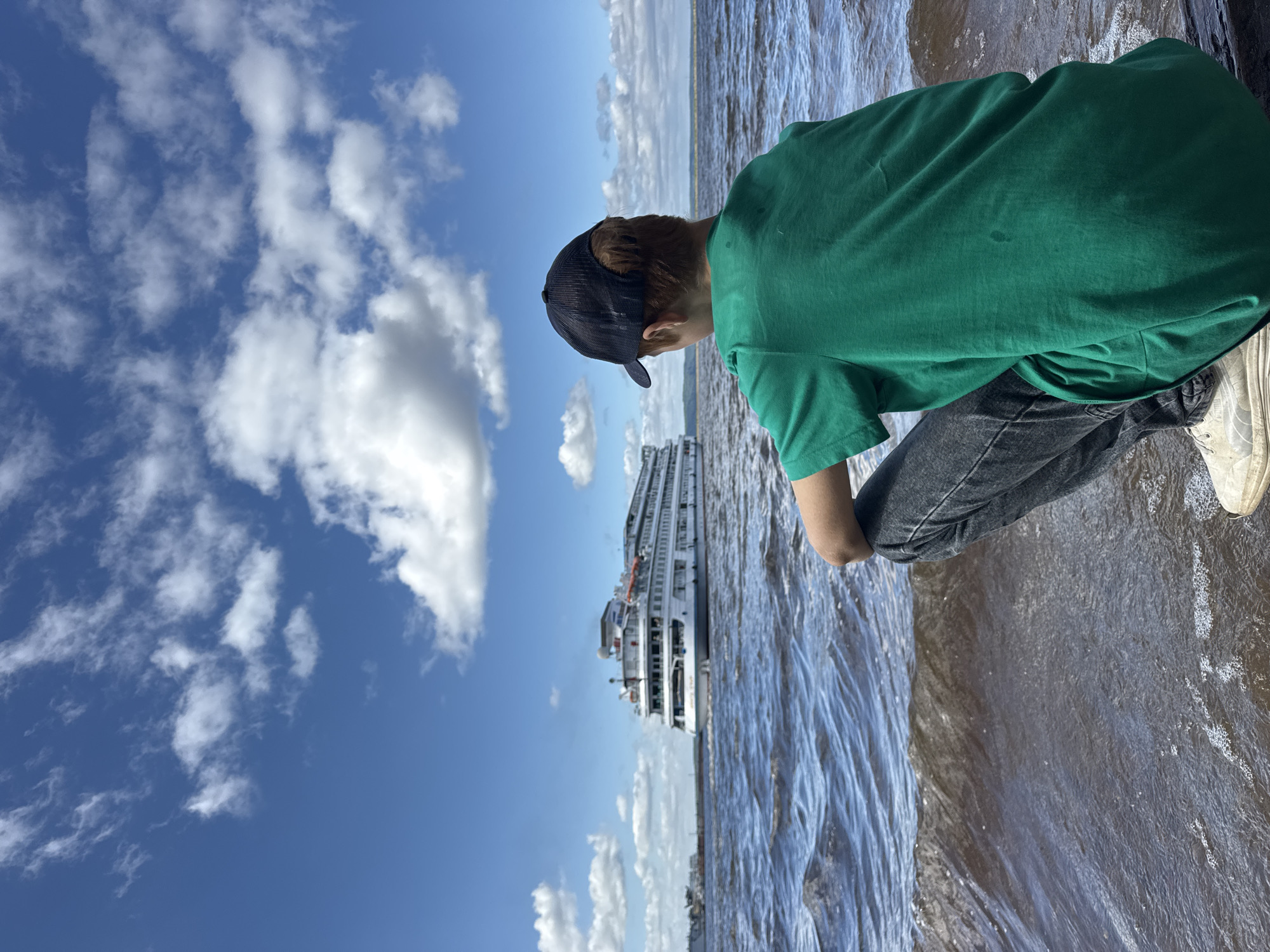 A young person wearing a green t-shirt and a black baseball cap is crouched down on a wooden surface, facing away from the camera, looking out at a large white passenger ship on a wide river. The water is dark and choppy, with small waves lapping at the edge of the wooden platform. The sky is a vibrant blue, dotted with fluffy white clouds. In the distance, on the far bank of the river, there is a faint line of trees. The scene appears to be a calm and pleasant day by the water. The image was likely taken in the city of Chaikovsky, Russia, given the presence of the passenger ship which is characteristic of river cruises on the Kama River in that region.