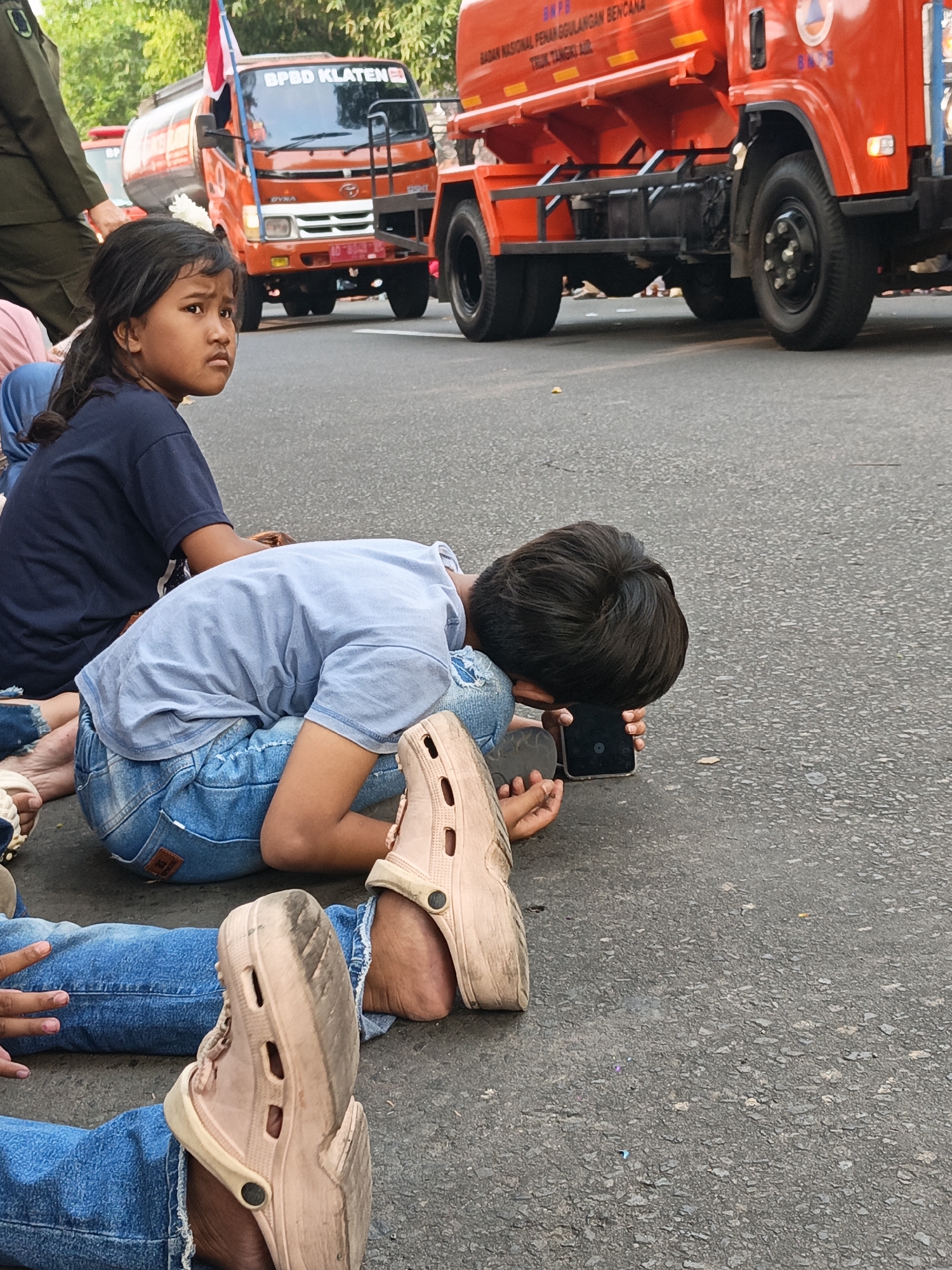 A young girl with dark hair pulled back in a ponytail sits on the asphalt in Gayamprit, Indonesia, looking to her left with a serious expression. She wears a navy blue t-shirt. To her right, a young boy with dark hair is curled up on the ground, resting his head on his knees and looking at a smartphone held in his hand. He is wearing a light blue t-shirt and denim jeans. His feet, clad in light pink Crocs, are visible in the foreground. Also in the foreground, another person's legs are visible, wearing blue jeans and one foot in a similar pink Croc. Behind the children, an orange truck with "BPBD KLATEN" written on its side is parked on the road, with a red and white flag attached to its roof. The truck appears to be a water tank truck, indicated by its cylindrical tank. The environment is an outdoor, paved surface, likely a street, with trees visible in the background.