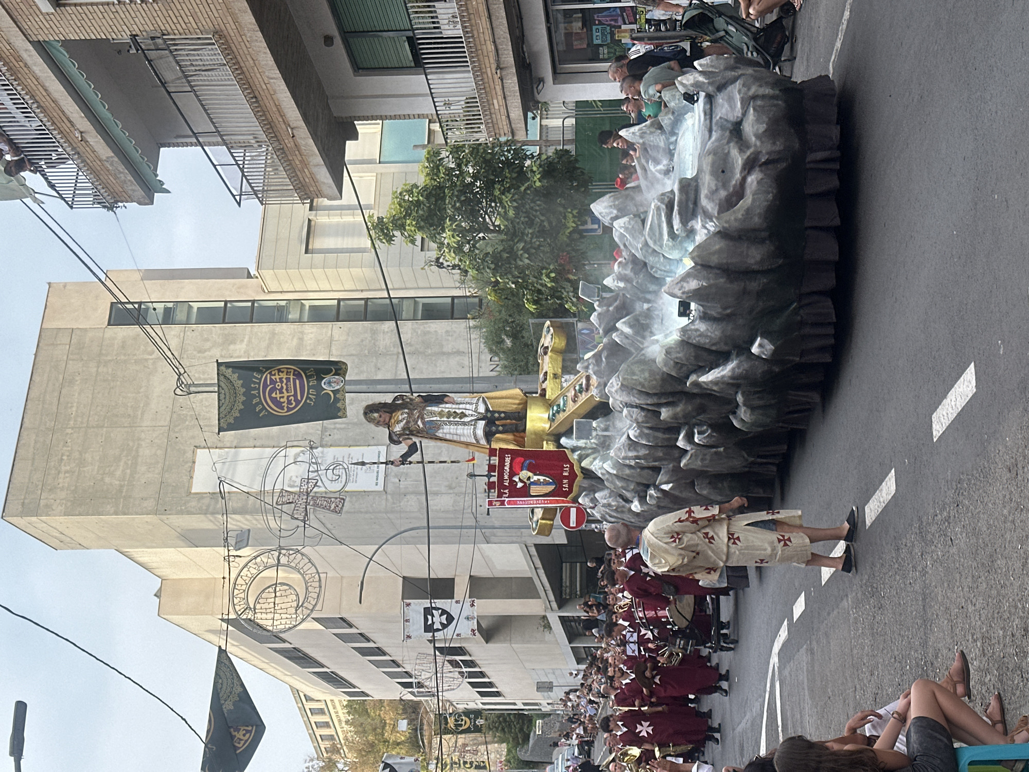 This image depicts a street parade in Alicante, Spain, likely during a festival or religious procession. A large float resembling a rocky, watery landscape with steam effects is prominent in the foreground. On top of the float stands a person in ornate armor, holding a spear. To the left of the float, a group of people in matching burgundy robes with white cross symbols are marching, some playing brass instruments, indicating they are part of a musical ensemble. Another individual in a white robe adorned with red crosses walks near the float. Spectators are visible on the sidewalks, watching the procession. In the background, buildings with balconies and banners hang from lampposts, suggesting an urban setting. The sky is overcast, and the lighting indicates it is daytime. The architectural style of the buildings and the general atmosphere suggest a Spanish city.