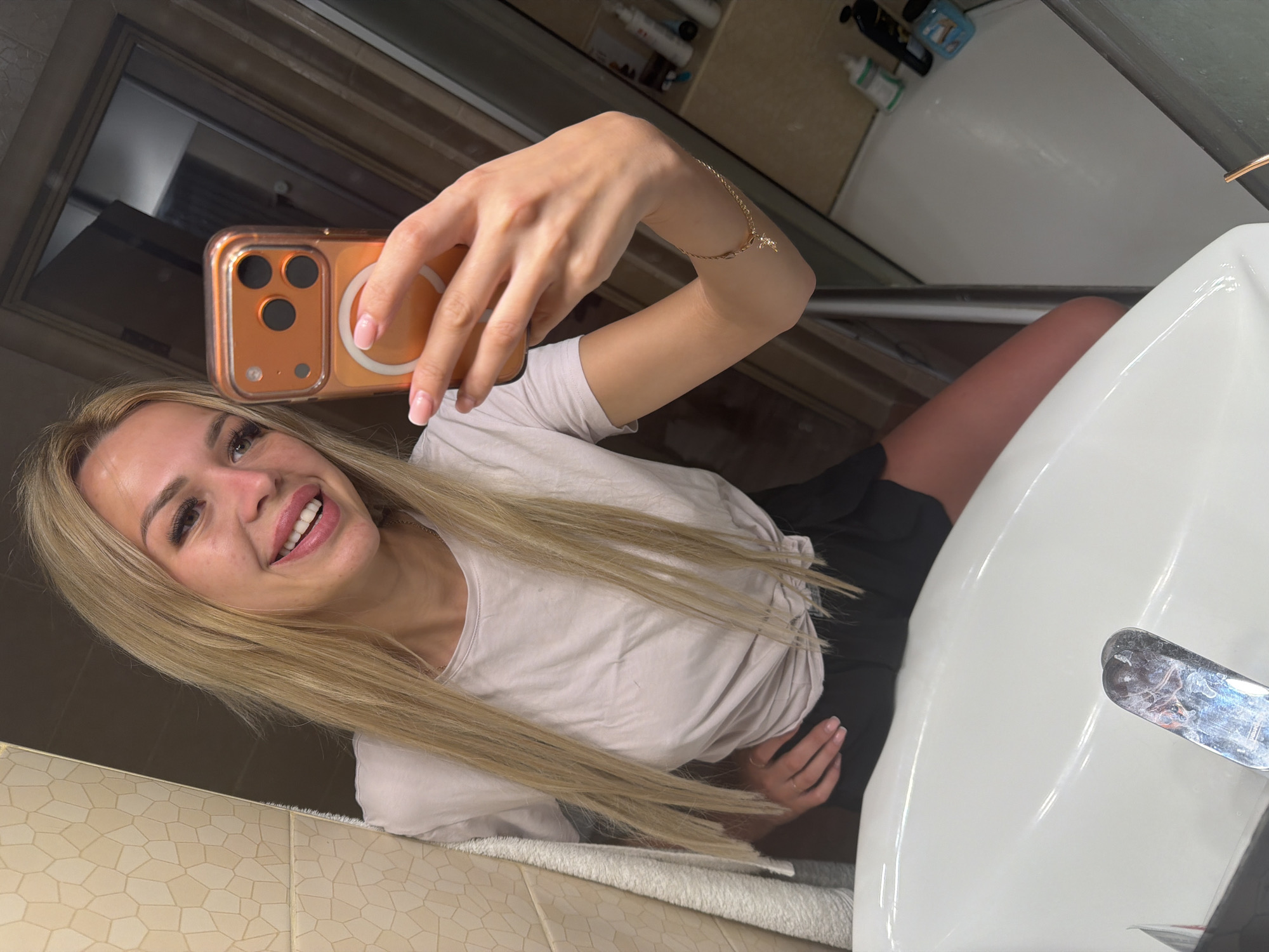 The image shows a young woman taking a selfie in a bathroom. She has long blonde hair, is wearing a light pink t-shirt, and appears to be smiling. She is holding an orange smartphone in her right hand, which is reflecting in the mirror. A bracelet with a small charm is visible on her left wrist. The background shows a tiled wall, a white sink, and what appears to be a bathtub. Bottles of toiletries are visible on a shelf behind the sink. The lighting suggests it is daytime. The image is taken from a low angle, looking up at the woman.