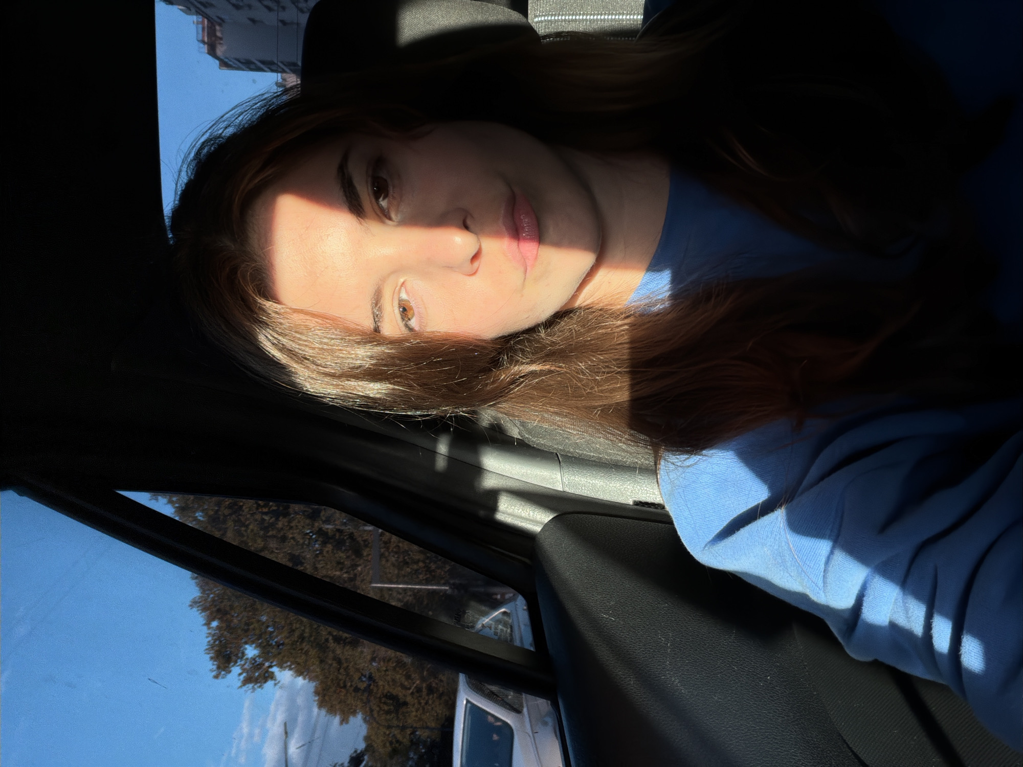 A young woman is sitting in the passenger seat of a vehicle, likely a car, with her face partially illuminated by sunlight. She has light skin, brown hair, and brown eyes. She is wearing a light blue sweatshirt. The interior of the car is visible, including the dashboard, window, and door panel. Outside the window, trees and a building can be seen under a blue sky, suggesting a sunny day. The location, city of Buenos Aires, Argentina, cannot be confirmed from the image alone.