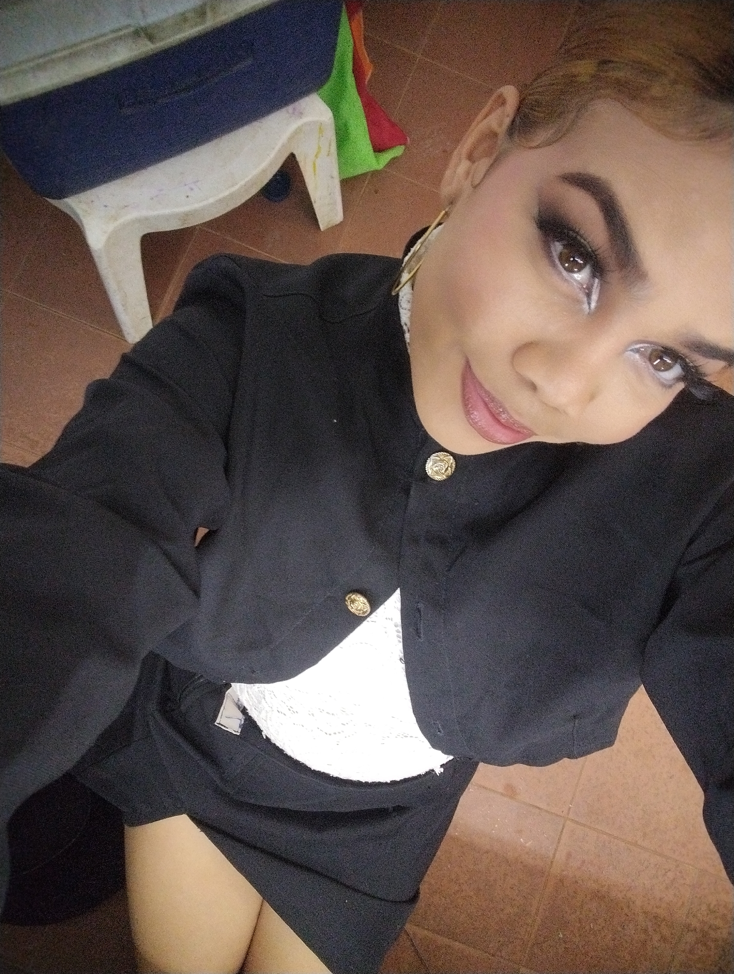 A young woman with elaborate eye makeup and light lipstick poses for a selfie in what appears to be a tiled room. She wears a black blazer over a white lace top and a black skirt. Gold-toned buttons adorn the blazer. Her hair is light brown and styled back. In the background, a white plastic stool and a blue and grey object, possibly a laundry hamper, are visible. The floor is tiled with brown squares. The image is a close-up and slightly angled shot, capturing the woman from the chest up. The overall setting suggests an indoor environment, potentially a home or a changing room. The location in Porlamar, Venezuela is an added detail, but without specific landmarks, it's not directly verifiable from the visual information alone.
