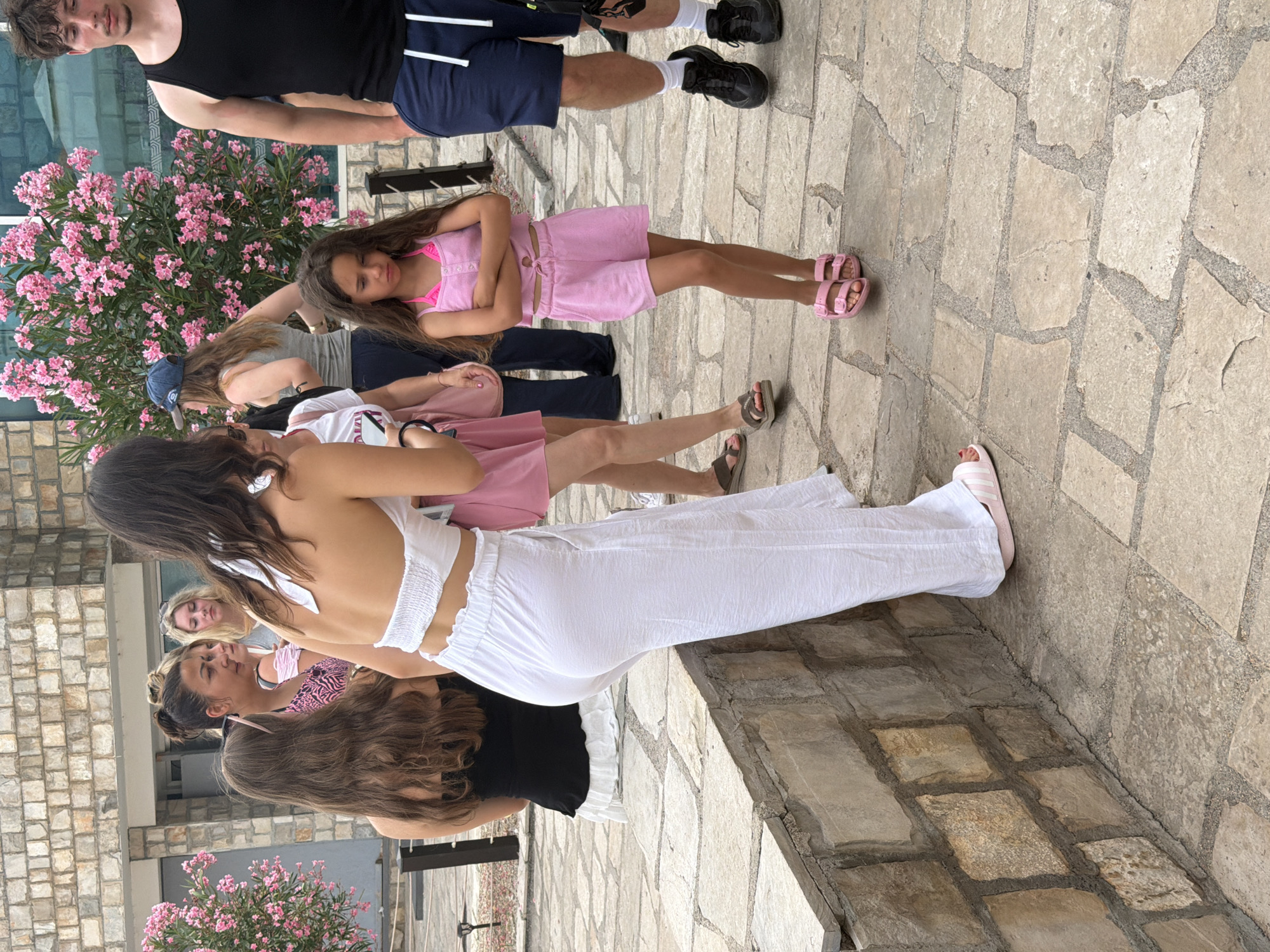 The image shows a group of people, appearing to be teenagers and young adults, gathered outdoors on a stone pathway. The setting is likely a tourist location, possibly in Budva, Montenegro, given the architecture and vegetation. In the background, a large bush with bright pink flowers is visible. Some of the people are wearing casual summer clothing, such as shorts, tank tops, and dresses. One individual in the foreground is wearing a white, ruffled crop top and flowing white pants, and another person is wearing a pink matching set consisting of a crop top and shorts. A young girl in a pink outfit is positioned slightly behind the group in the foreground. The lighting suggests it is daytime, possibly late afternoon or early evening, with soft shadows. The overall mood seems relaxed and informal, with people interacting and socializing. There is no visible text in the image. The ground is paved with irregular stone tiles.