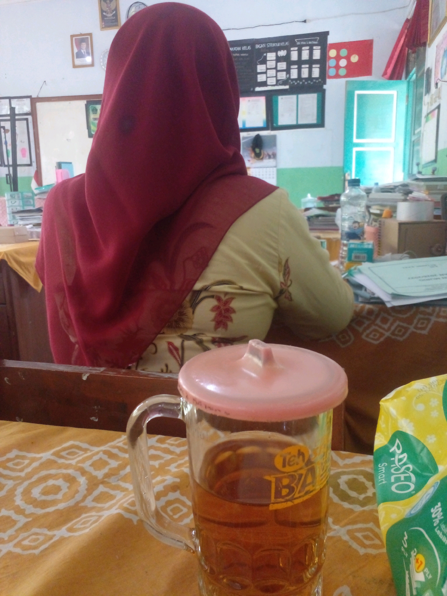 An overhead medium shot shows a woman from the back wearing a dark red hijab and a light yellow blouse with red floral patterns. She is seated at a wooden desk. In the foreground, a clear glass mug filled with amber-colored liquid, likely tea, rests on an orange and white patterned cloth. The mug has a pink lid. To the right of the mug is a partially visible yellow and green package of Paseo Smart wet wipes. The background reveals a classroom setting with educational charts on the wall, a blue-green door, and stacks of papers and books on a desk. The overall impression is of a student or teacher taking a break in a classroom in Banyuputih, Indonesia.