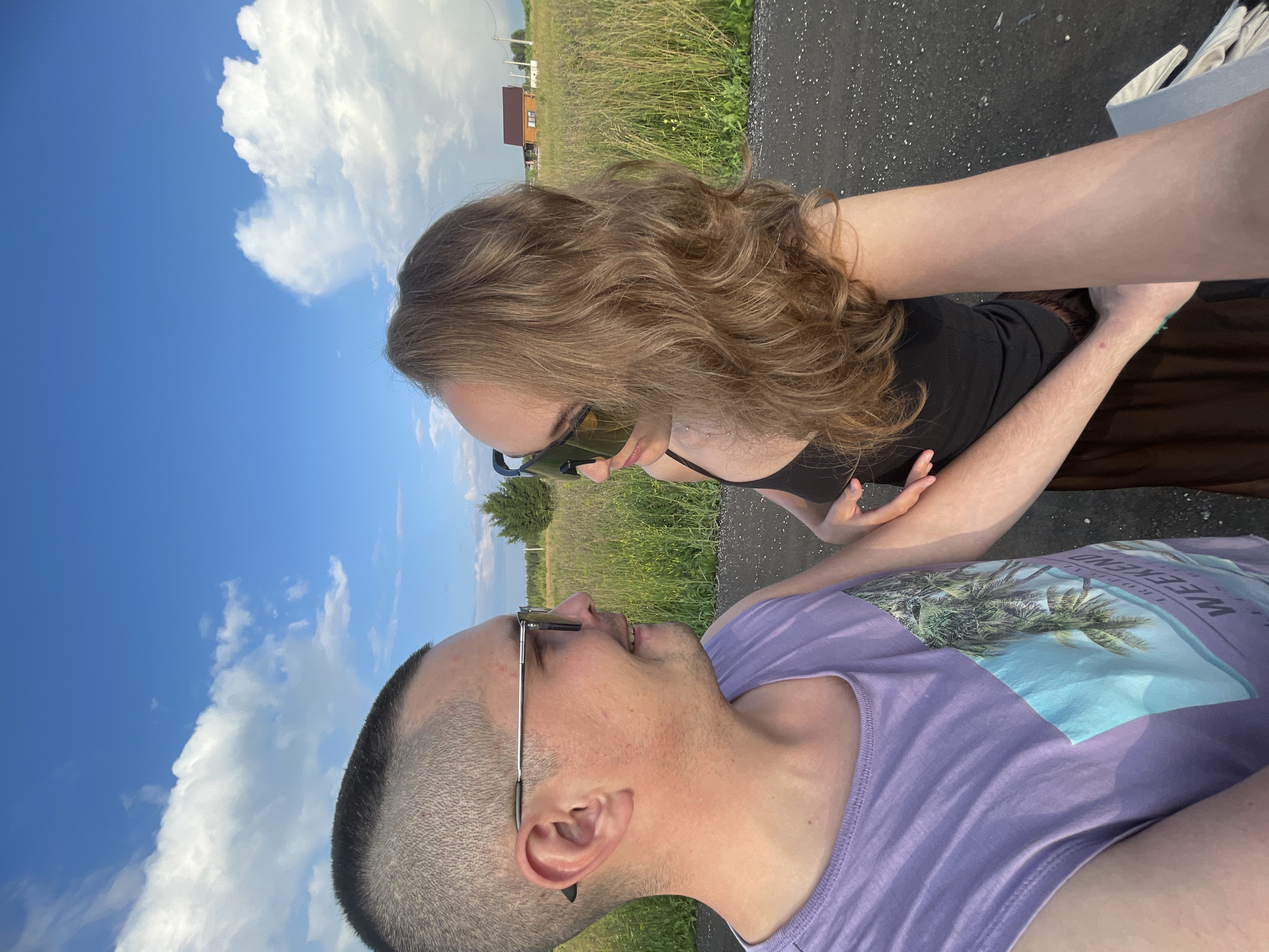 An outdoor, close-up, selfie-style photograph depicts two individuals, likely a couple, against a backdrop of a rural landscape under a bright blue sky with scattered white clouds. The individual on the left, a person with a shaved head and wearing a light purple tank top with a palm tree graphic, is looking to their right, smiling. They are wearing silver-rimmed sunglasses. The individual on the right, with long, wavy blonde hair, is wearing a black tank top and large, dark sunglasses. They are looking towards the left. Their arms are intertwined. The background shows a field of tall, green grass and some trees, with a small wooden house visible in the distance. The ground in the foreground is a dark, paved surface, possibly a road. The scene appears to be set in the village of Podshivalovo, Russia, given the context of the request.