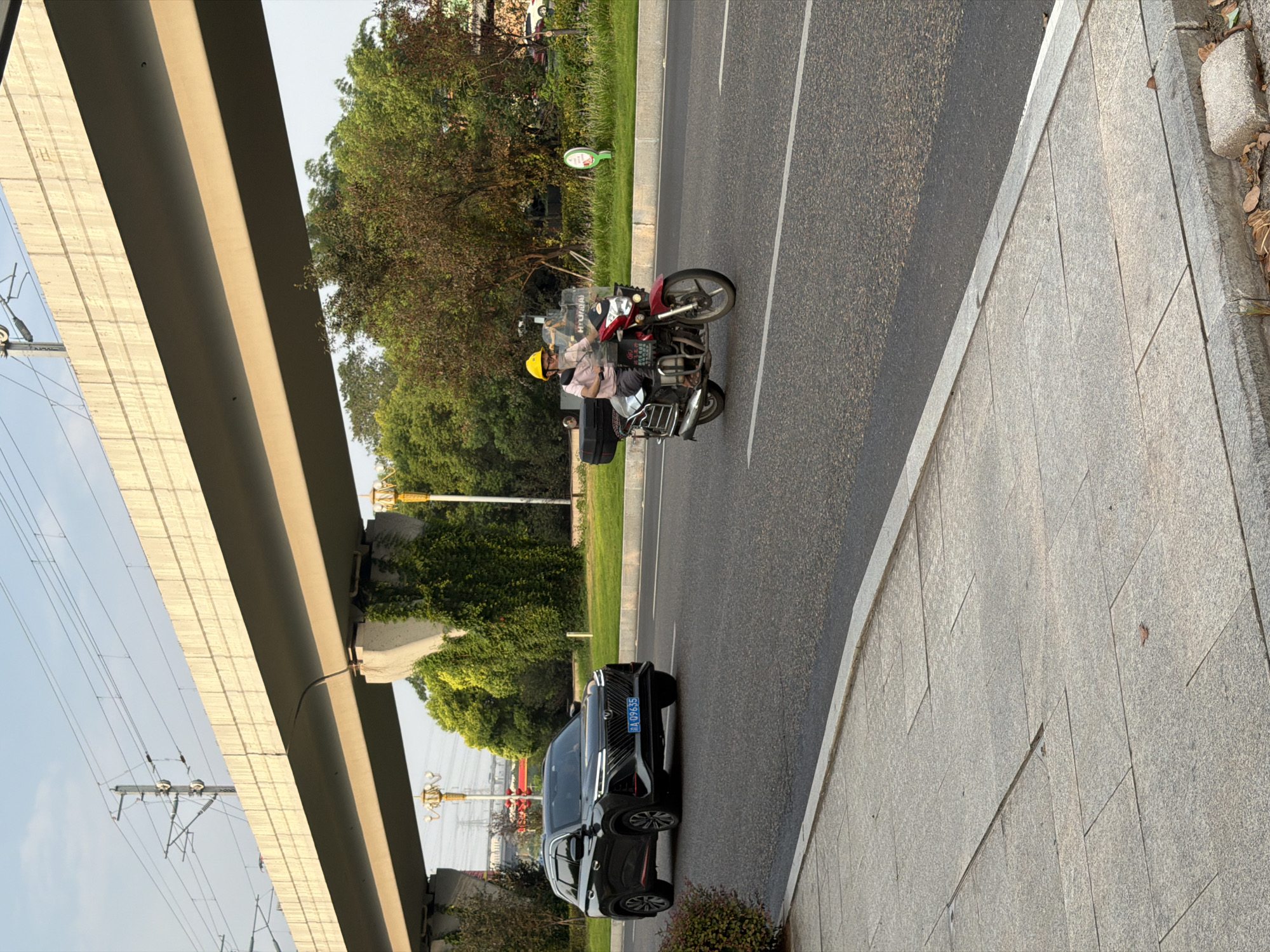 In Jiangjin, China, on a street beneath an elevated highway, a dark-colored SUV is traveling on the right lane of a two-lane asphalt road. A person wearing a yellow helmet and a light pink shirt is riding a red and silver motorcycle in the left lane, moving in the same direction as the SUV. The motorcycle has a black storage box mounted on the rear. The road is bordered by a sidewalk made of gray granite slabs. Beyond the road, there are green trees, some with red leaves, and vegetation growing on concrete pillars supporting the highway. The sky is clear and light blue.
