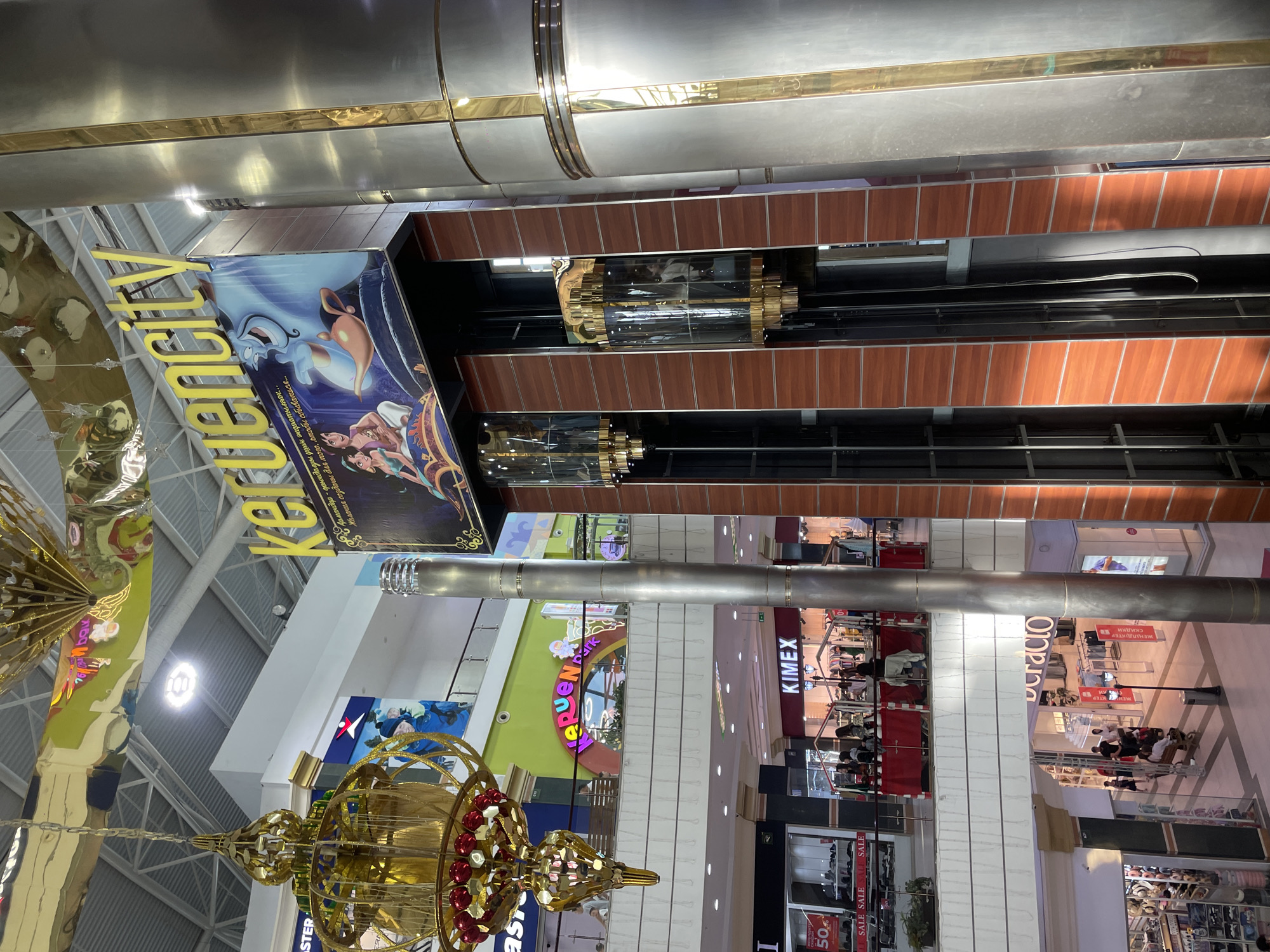This is an indoor shot of a multi-story shopping mall in Aktobe, Kazakhstan. The perspective is looking upwards towards the ceiling, revealing the architectural details of the mall's interior. Dominating the scene is a large banner featuring characters from Disney's Aladdin, with the text "Keruencity" prominently displayed in yellow lettering. The banner is a vibrant blue and purple, and it appears to be advertising a promotion or event related to the movie.
Below the banner, the mall's interior is visible, showing multiple levels with shops and escalators. The architecture features a mix of modern materials like glass and metal, contrasted with wood-paneled accents. There are several large, cylindrical metal structures running vertically through the mall, possibly part of the ventilation system or decorative elements. On the upper levels, shop fronts with names like "KIMEX" and "Doracio" are visible. Some shop windows display clothing and accessories, with "SALE" signs indicating discounts.
On the lower levels, there are more shops and people browsing. A large, ornate golden chandelier with red accents hangs in the foreground, adding a touch of grandeur to the space. The lighting suggests it is daytime, as natural light filters in from high windows, though the interior also features artificial lighting. The overall impression is that of a busy, modern shopping environment. The text on the banner and within the shops indicates the location as Aktobe, Kazakhstan.