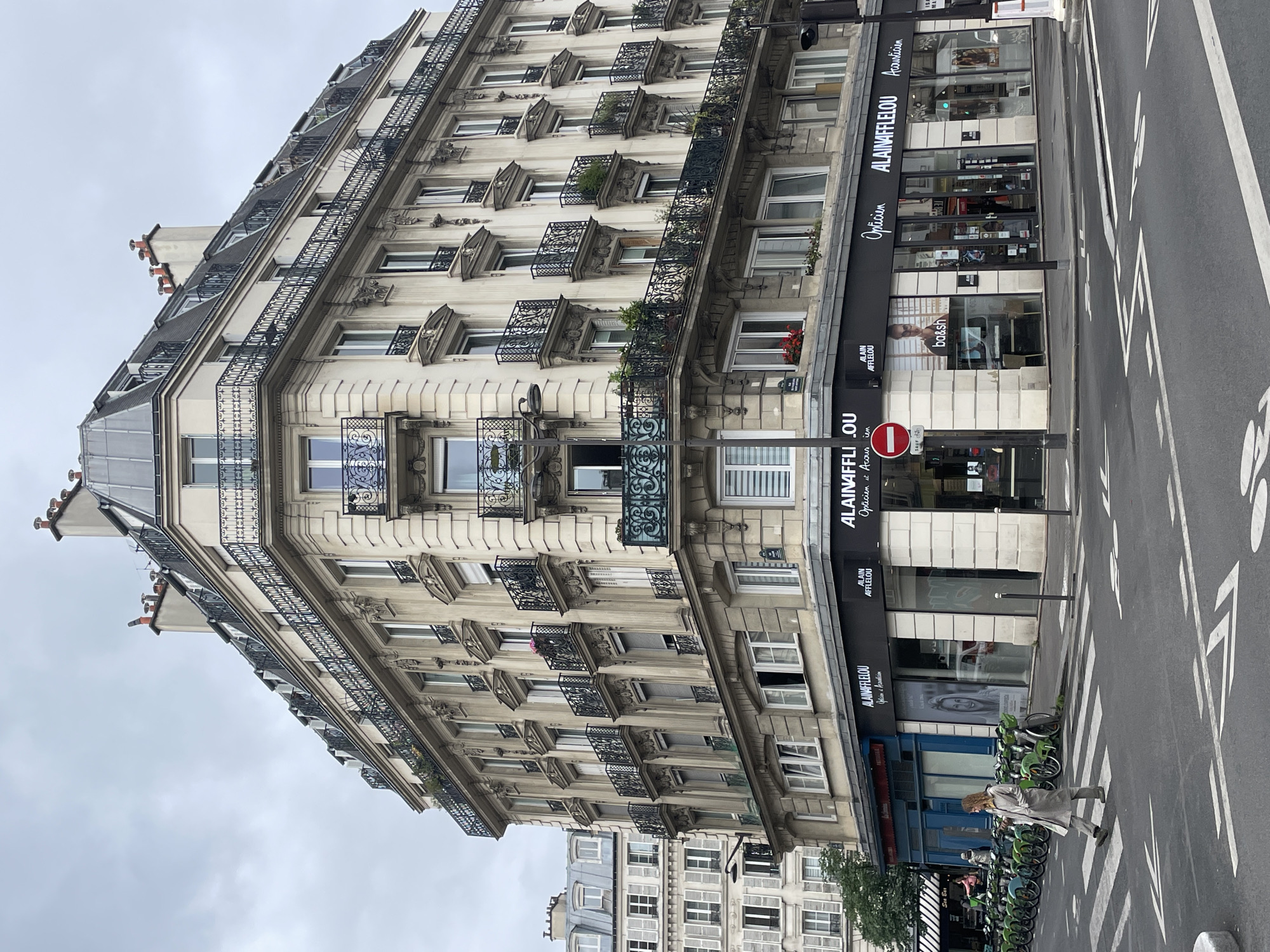 This image depicts an urban street scene in Paris, France, dominated by a classic Haussmann-era building. The prominent, multi-story structure features a light-colored stone facade with numerous windows, each adorned with ornate wrought-iron balconies and intricate decorative carvings. The building is topped by a grey mansard roof with dormer windows and several chimneys, indicative of Parisian architecture.
At street level, commercial establishments occupy the ground floor. A prominent shop is "ALAIN AFFLELOU," identified as an "Opticien et Acousticien" (Optician and Acoustician) on its black signage, which appears multiple times. One of the display windows features a poster for the brand "ba&sh." A red and white "no entry" traffic sign is visible near the storefront. The street in the foreground is an asphalt road with white markings, including a crosswalk. Along the sidewalk, a row of shared bicycles, appearing to be Lime bikes, is parked. One person, dressed in a light-colored coat, is seen walking on the sidewalk beside the bicycles.
The scene takes place during the daytime, under an overcast, grey sky. The distinctive architecture, the French text on the storefronts, and the presence of shared bicycles common in major European cities strongly indicate the location. Other buildings with similar architectural styles are visible in the background.