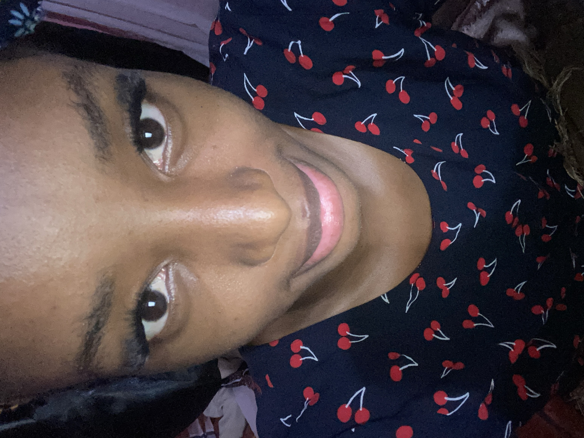 A close-up, high-angle selfie captures a young adult individual from the forehead to the upper chest, looking directly at the camera. The subject has dark brown skin, prominent dark brown eyes with distinct white rectangular reflections in the pupils, indicating a light source directly above or in front. Dark, full eyelashes and well-defined, somewhat bushy dark eyebrows frame the eyes. The nose has a broad bridge. The mouth is slightly open, revealing a subtle, light pink-tinged smile with the tip of the tongue just visible. The lips appear moist. The individual wears a dark navy or black round-neck top patterned with multiple small red cherries, each featuring a white stem and leaf outline. A portion of a dark headphone or headset band is visible near the subject's left ear (viewer's left). The background is out of focus. To the viewer's left, a dark, indistinct mass suggests fabric or furniture. To the viewer's right, blurry vertical stripes in shades of pink, purple, and white are visible, consistent with curtains or soft furnishings. The lighting is even, illuminating the subject's face, which exhibits a slight sheen on the forehead and nose. The image is stated to be captured in the city of Jos, Nigeria.