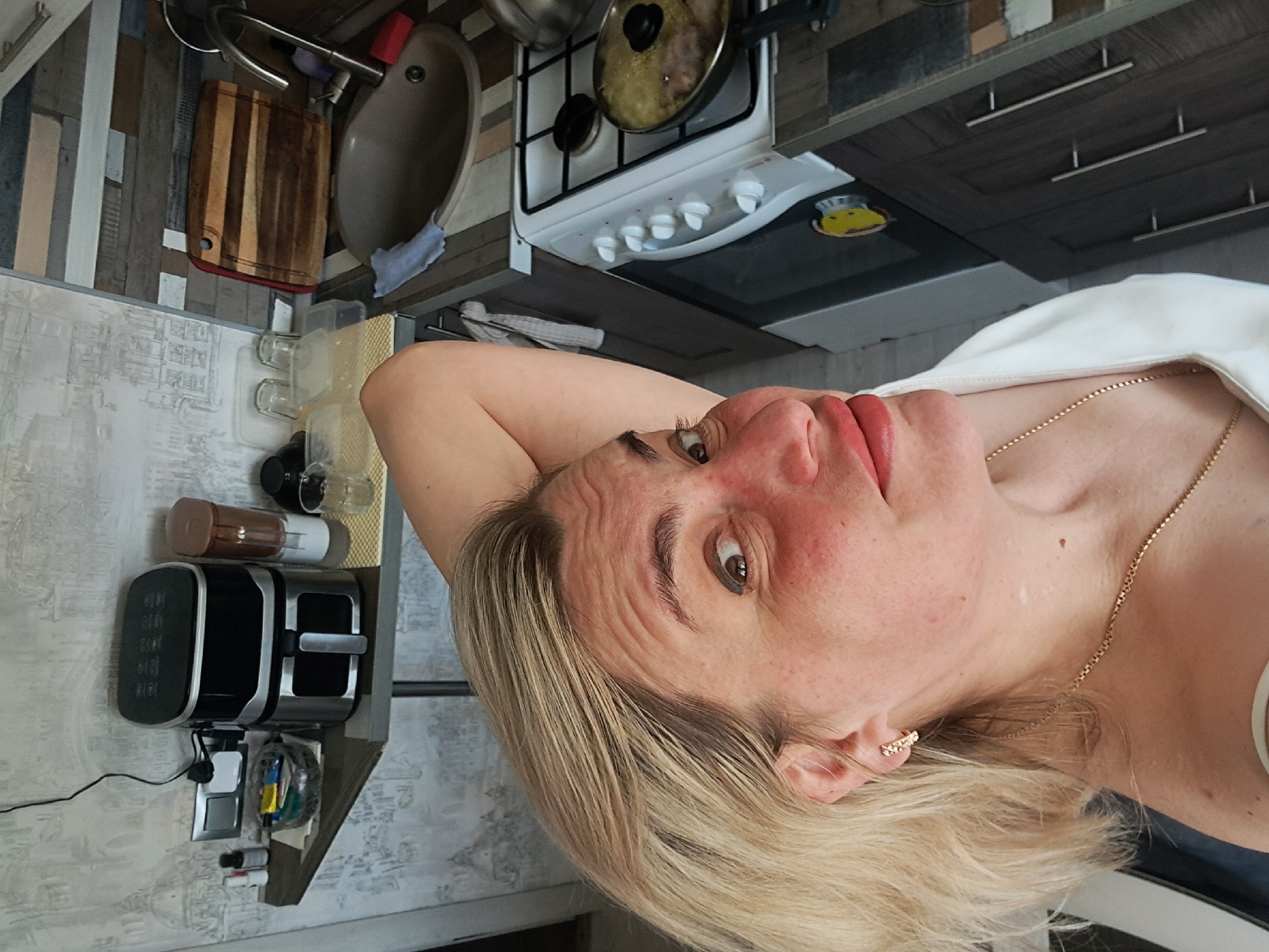 A woman with fair skin and blonde hair is positioned in the foreground, appearing to be indoors. She is wearing a gold necklace and has a white garment draped over her shoulder. Her face is angled towards the camera.
In the background, there is a kitchen setting. A white gas stove with a pot on one burner and a range hood above it is visible. To the left of the stove, there is a sink and a cutting board hanging. Beneath these is a kitchen wall-mounted appliance. The walls are painted in a patterned motif. A cabinet is partially visible in the top right corner of the image.
The location is Elektrostal, Russia, indicated by possible metadata or source information, which suggests a domestic, residential setting. The focus is on the woman in the foreground, but the background establishes a kitchen environment.