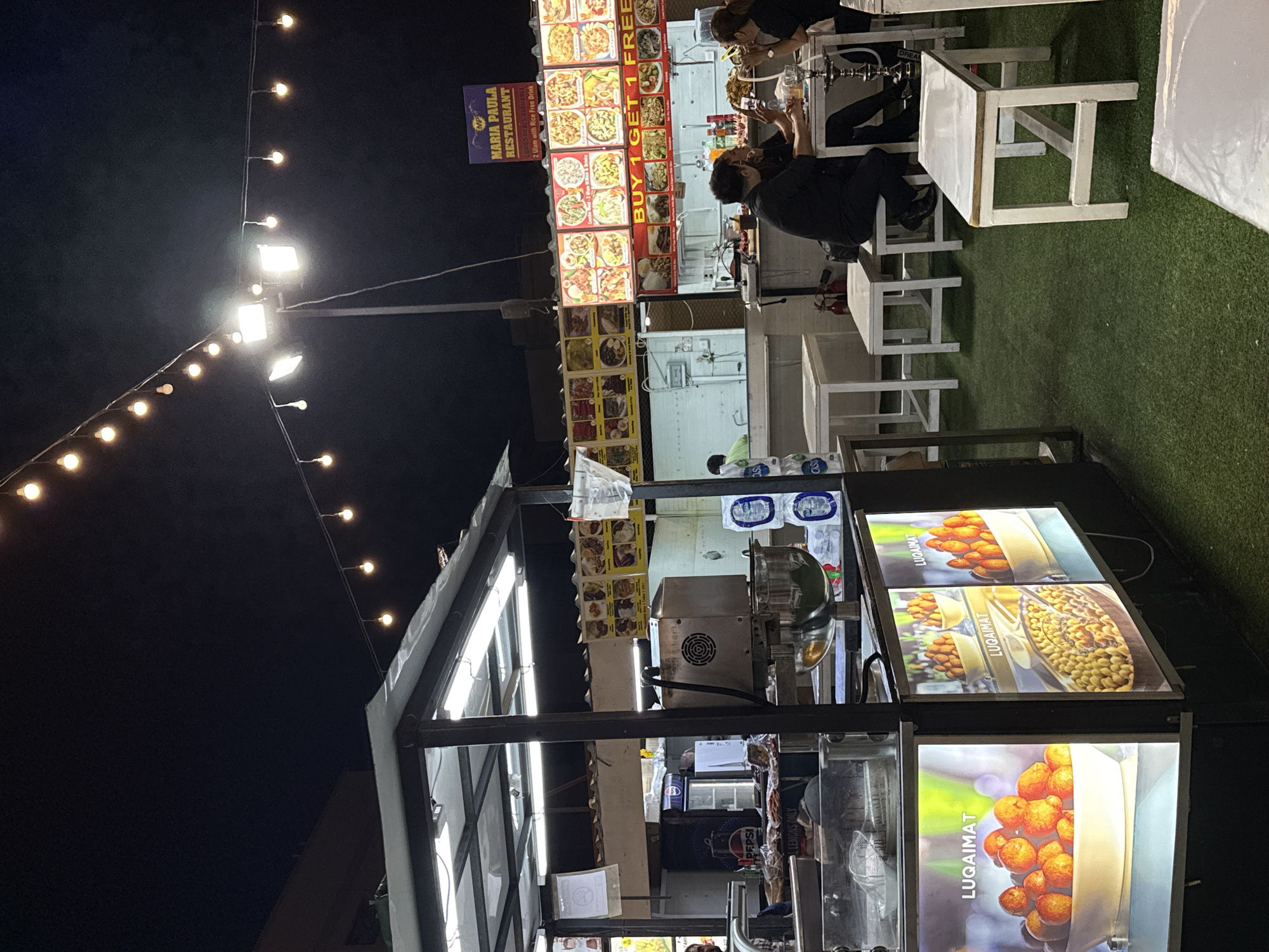The image shows an outdoor food establishment in Dubai, United Arab Emirates, at night. The brightly lit "Maria Paula Restaurant" sign and "Buy 1 Get 1 Free" offer are visible on the restaurant facade. People are seated at white tables, some using a hookah.
A food stand in the foreground advertises "Luqaimat", a local dessert. The stand has illuminated display panels. A metal food-making machine is also visible. The lighting includes strings of lights and bright spotlights.
