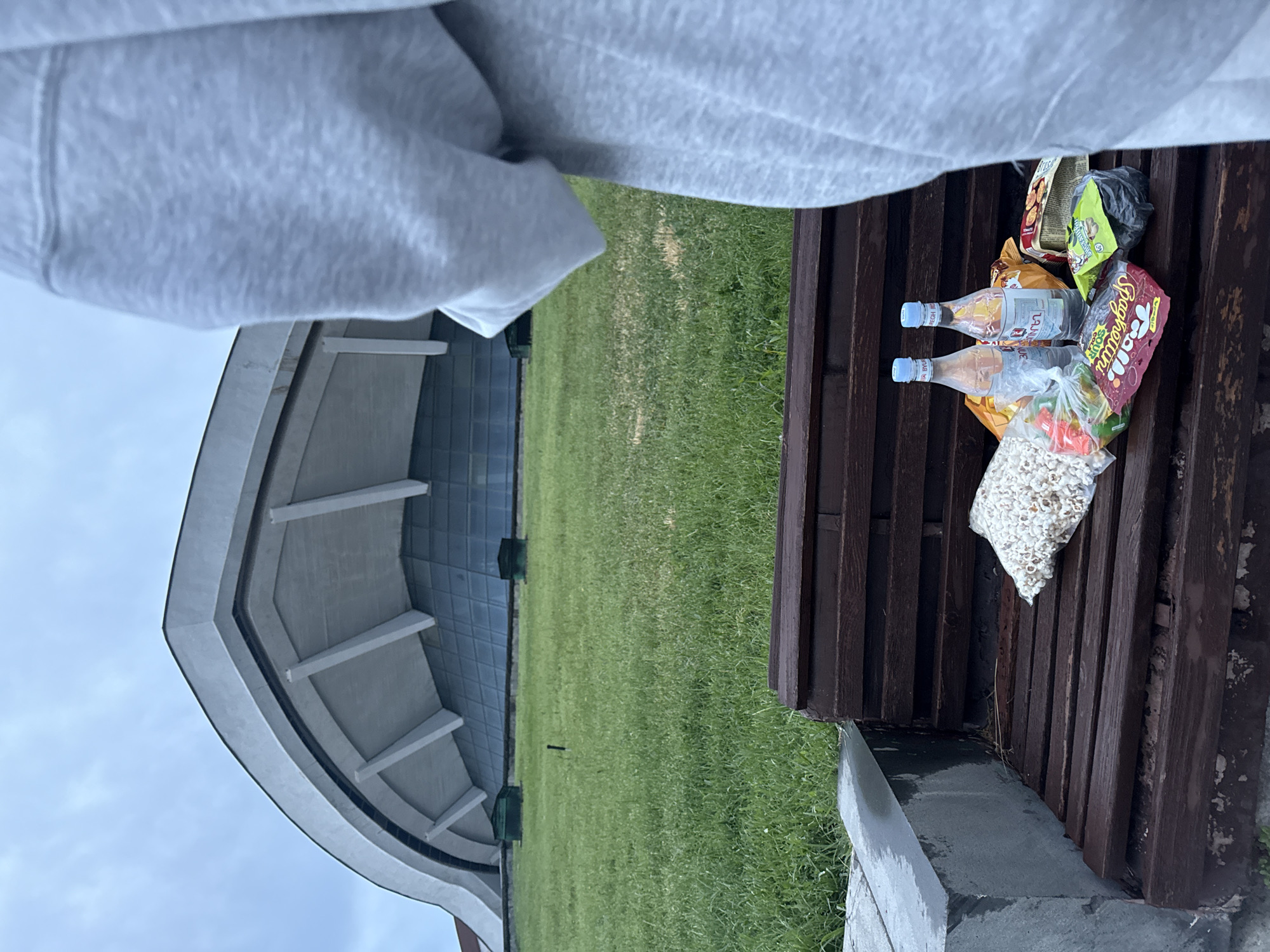 A horizontal image shows a bench laden with snacks and drinks in front of a modern building amidst a green field, likely in Yerevan, Armenia. Two clear plastic bottles with blue caps, possibly mineral water or lemonade from "Byuregh", sit among assorted bags of chips, candy, and popcorn on the dark brown wooden slats of the bench. One bag is labeled "Spaghettini Soup". The bench is situated near a concrete curb, with a lush green field stretching toward a contemporary structure in the background. The architecture features a curving roof and what appears to be a glass facade. A portion of someone's gray hooded sweatshirt is visible in the upper left. The sky is overcast, creating soft, diffused lighting.