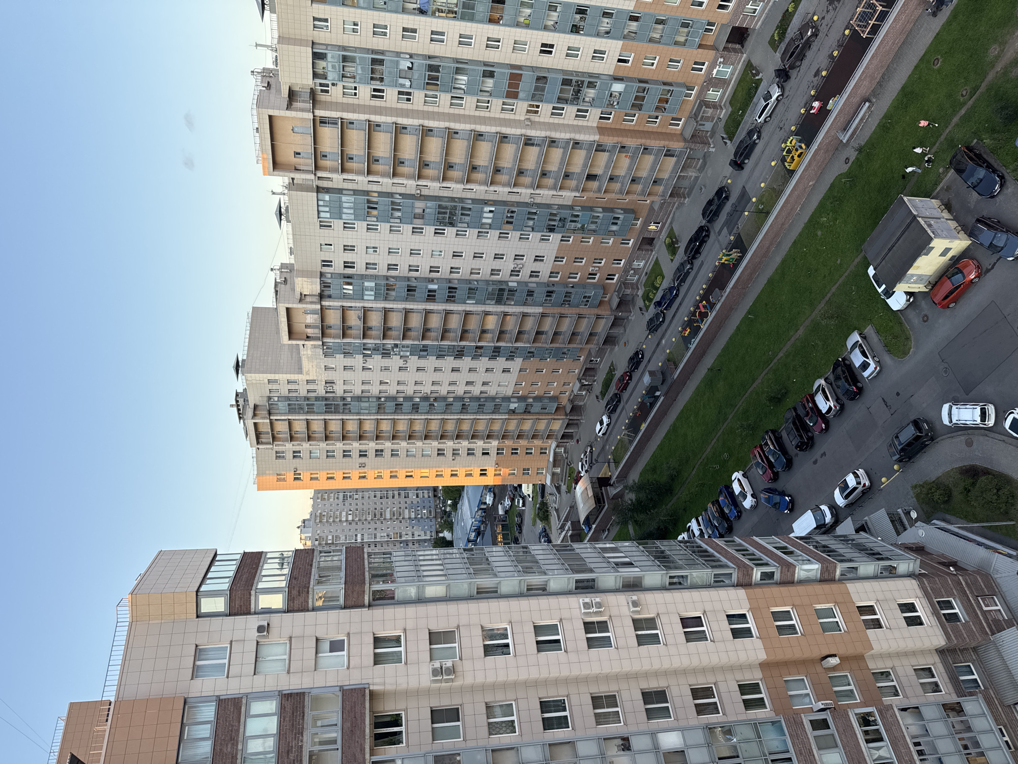 An aerial, high-angle view captures a city street lined with multi-story apartment buildings. The buildings are modern, constructed with light-colored panels and featuring numerous balconies and windows. Cars are parked along the street and in a designated lot. A green, grassy area separates the street from the buildings, with a brick wall along its edge. A small yellow shed-like structure is present near the grassy area. The sky above is a clear, pale blue.