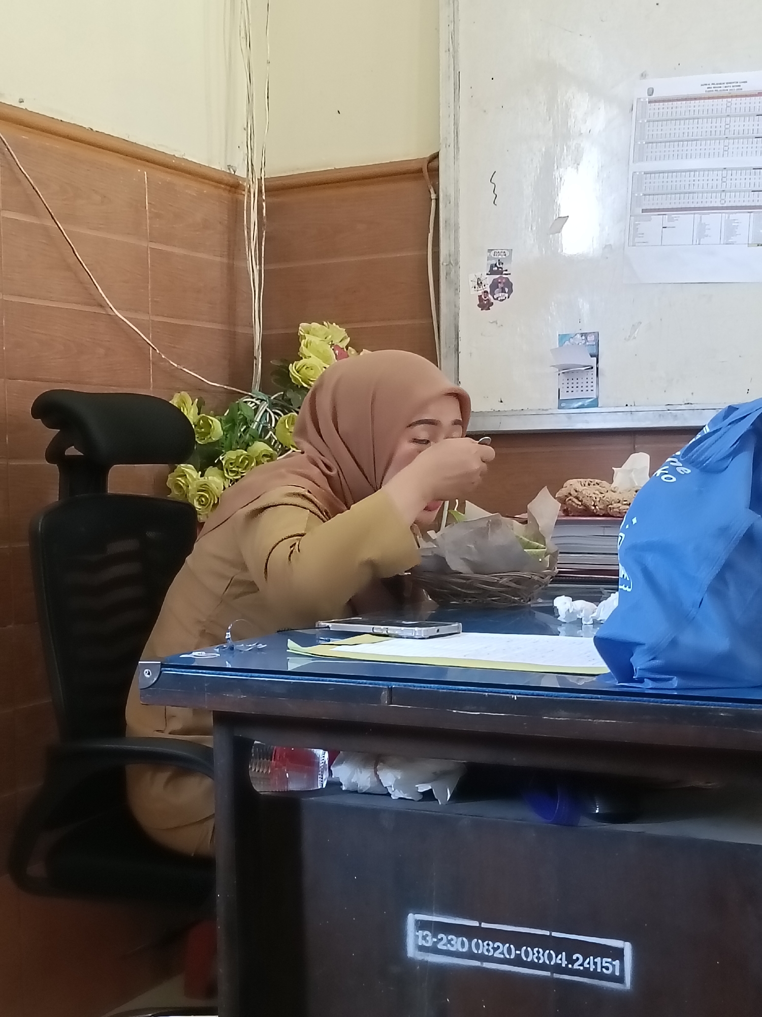 The image captures a medium shot of a woman wearing a brown hijab and a brown uniform, seated at a dark blue desk in what appears to be an office environment. She is actively eating from a woven basket containing food, holding a spoon to her mouth. To her right, a bright blue reusable shopping bag is prominently displayed. The desk surface is cluttered with papers, a smartphone, and crumpled white tissues. Behind the woman, a black office chair is visible, and further in the background, a wall with wooden paneling and a white noticeboard adorns the space. The noticeboard displays various documents, calendars, and stickers. A bouquet of artificial yellow roses rests on a shelf or surface behind the woman, adding a touch of decoration to the room. The overall setting suggests a workspace where a brief personal moment, such as a meal, is being captured. The location is identified as Kediri City, Indonesia.