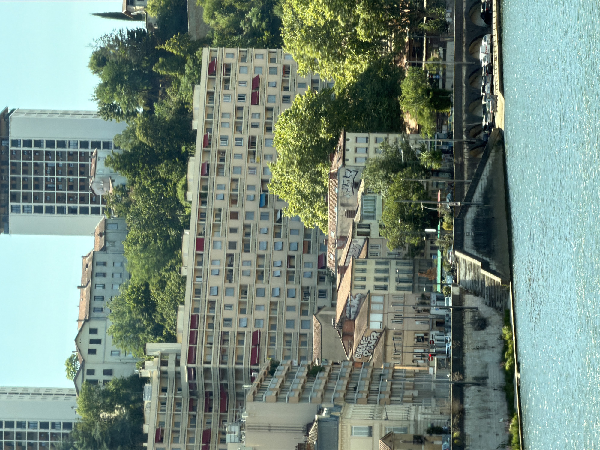 The image depicts a view of the Rhône River in Lyon, France, looking towards the Quai Saint-Vincent. In the foreground, the bright blue, slightly rippled water of the river dominates the lower portion of the frame. Along the riverbank, a concrete retaining wall with visible staining and texture is present, featuring a set of stone stairs leading up to a street level.
Behind the retaining wall, a row of buildings rises, characterized by their light-colored facades and repetitive window patterns. The buildings appear to be a mix of older, lower-rise structures with tiled roofs and more modern, multi-story apartment blocks. One prominent building is a large, cream-colored apartment complex with numerous balconies, many of which are adorned with red awnings. Further back, a tall, slender white building with a grid-like facade of windows stands out against the clear blue sky, likely another apartment building or office tower.
Vegetation, primarily lush green trees, is visible interspersed between and behind the buildings, suggesting a hilly or terraced urban landscape. Some of these trees are quite large, with dense foliage. The overall impression is of a dense urban environment with significant architectural development, softened by abundant greenery. The lighting suggests it is daytime, with the sun casting bright illumination.