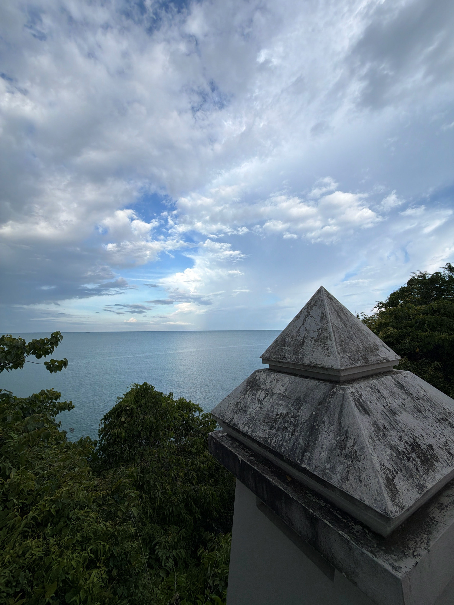 The image captures a serene view of the ocean and sky from a vantage point in Ko Samui District, Thailand. In the foreground, a weathered, grey, pyramid-shaped concrete structure, likely part of an outdoor decoration or architectural element, sits on a wall. Lush green foliage of trees and branches frames the scene from the left and right. The middle ground is dominated by the calm, blue expanse of the ocean meeting a hazy horizon. Above, a dramatic sky filled with a mix of white and grey clouds is partially illuminated by sunlight, with patches of blue sky peeking through. The overall atmosphere is peaceful and natural.