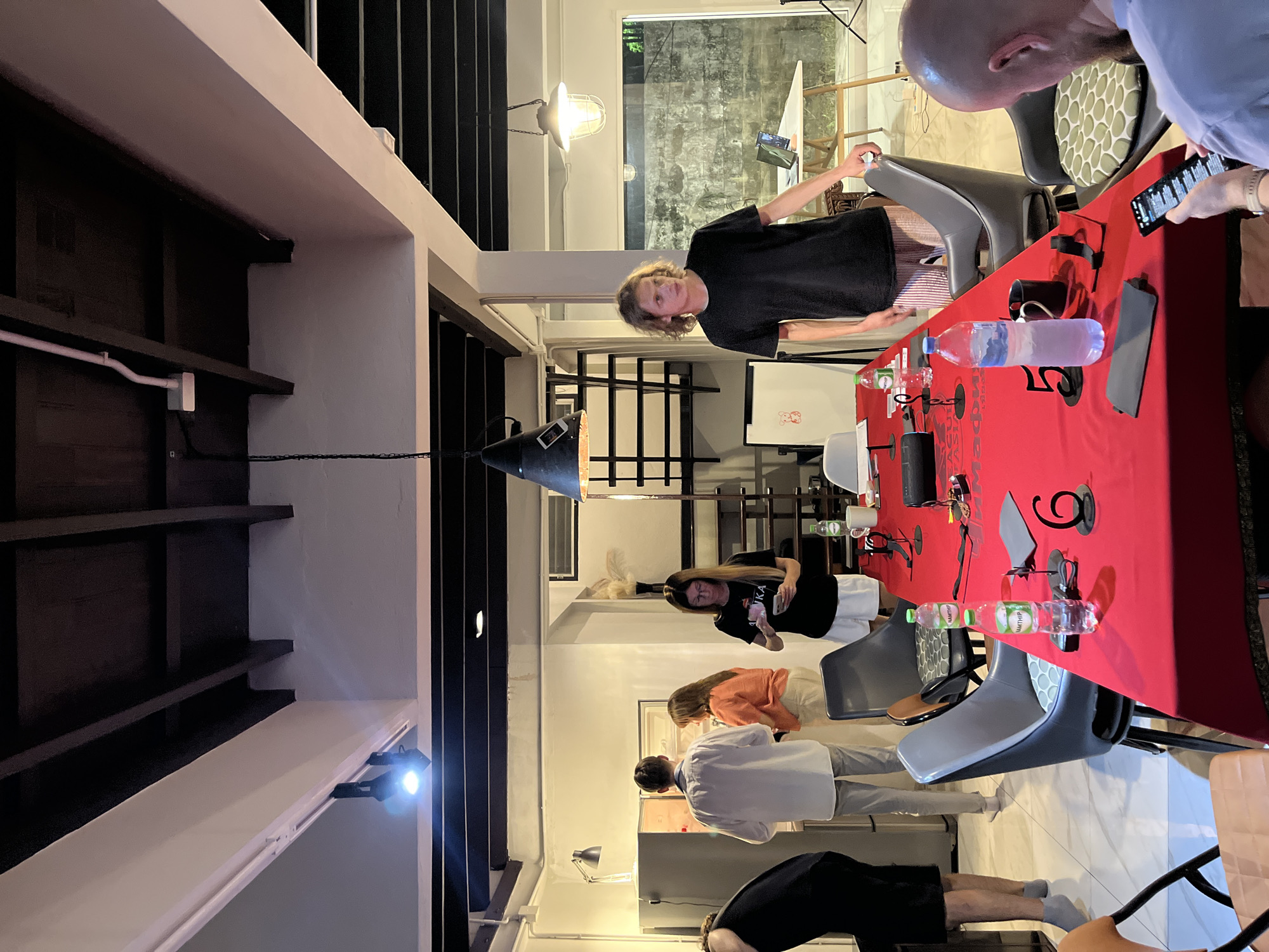 The image depicts a group of people gathered around a long table covered with a red tablecloth. The setting appears to be indoors, possibly a modern eatery or event space, with exposed beams and industrial-style lighting. The orientation of the image is sideways, making it appear as if the scene is on a wall.
There are at least six individuals visible, engaged in various activities. One person, with long blonde hair, wearing a black t-shirt, is looking towards the camera with a slight smile. Another person, with short, bald head, is looking down at a smartphone. Others are seen interacting with each other or looking at their phones. On the red table, there are several objects, including bottles of water, a coffee mug, and what appears to be a remote control. Text is visible on the water bottles, but it is difficult to decipher clearly.
The lighting suggests it might be evening or a dimly lit indoor space. The overall atmosphere seems casual and social, possibly a gathering or a work session. The presence of chairs and a table implies a shared activity or meal. The location context of Chiang Mai City Municipality, Thailand, is noted, though no specific visual cues directly confirm this in the image itself.