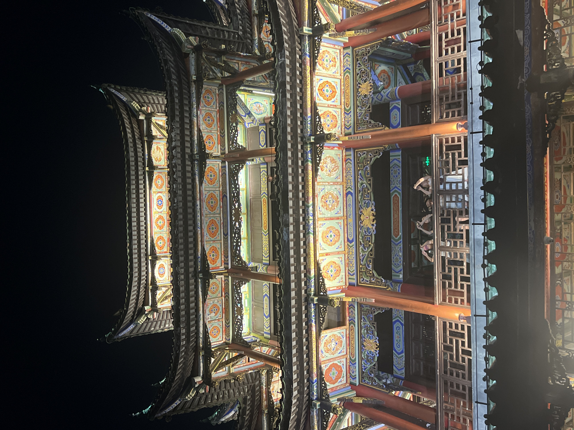 The image shows a multi-story traditional Chinese building at night, illuminated by warm lights. The architecture features layered, curved roofs with ornate decorative elements in red, orange, blue, and gold. Pillars painted red and decorated with gold motifs support the structure. Balconies with intricate wooden railings are visible on the upper levels. Several people, seen from the chest up, are standing on the balcony of the second story, some looking at their phones. A green illuminated sign is visible on the second story. The background is dark, indicating it is nighttime. The building is located in Liangjiang, China.
