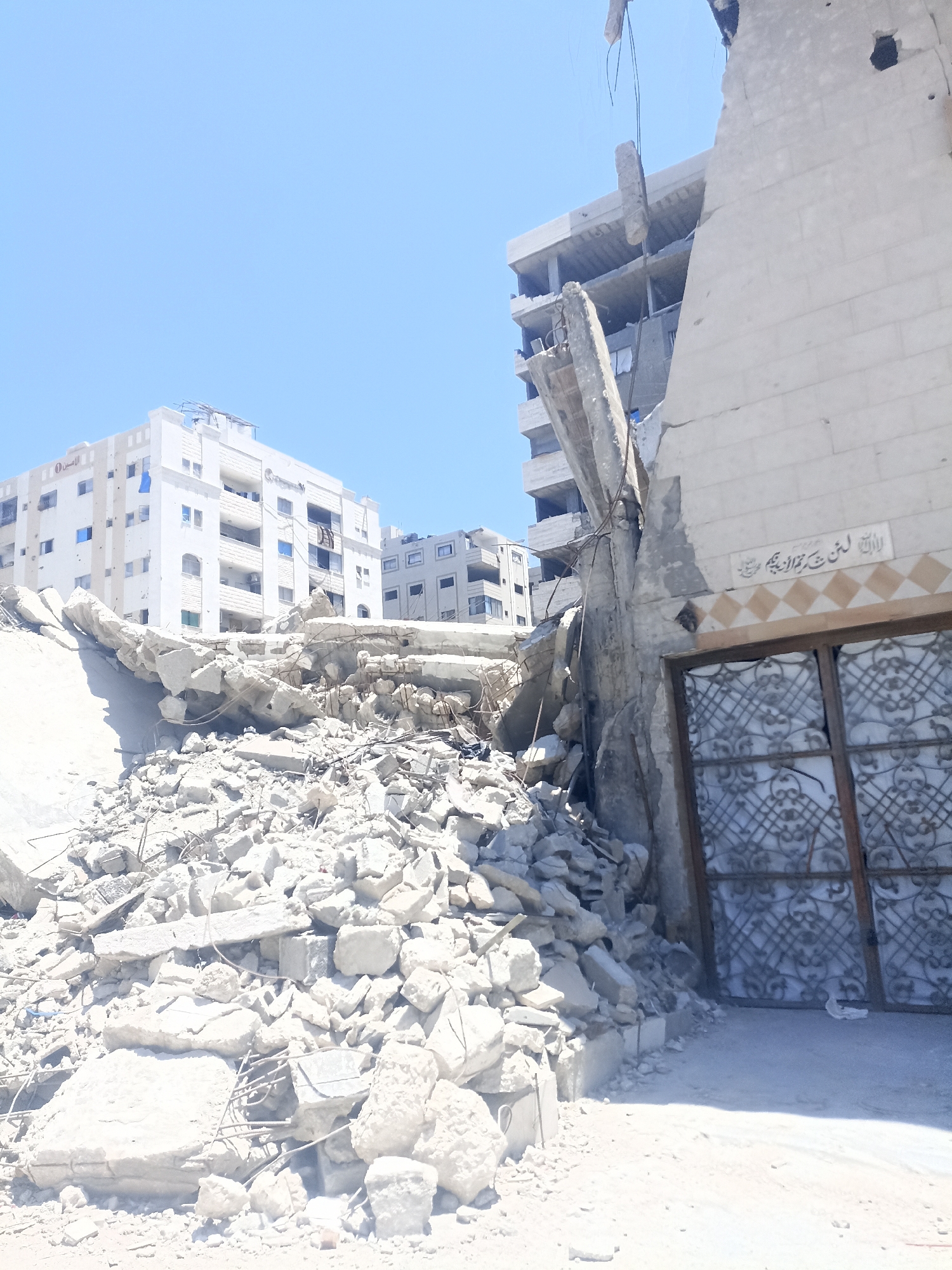 The image captures a scene of extensive destruction in the city of Gaza, Palestinian Territories, under a clear blue sky. In the foreground, a massive pile of rubble, composed of concrete debris, rebar, and other building materials, dominates the frame. These ruins suggest a recent catastrophic event, such as an airstrike or bombardment.
Behind the rubble, the skeletal remains of a multi-story building are visible. A large concrete pillar, fractured and leaning precariously, stands as a stark testament to the structural damage. Partially visible on the right side of the frame is another damaged building with a light-colored stone facade. Above a wooden-framed ornate metal gate on this building, Arabic calligraphy is inscribed on the wall. Further in the background, intact and partially damaged apartment buildings stand, their white and gray exteriors contrasting with the surrounding devastation. The scene is devoid of people, emphasizing the emptiness and aftermath of the destruction. The overall environment is dusty and chaotic, indicative of a conflict zone.