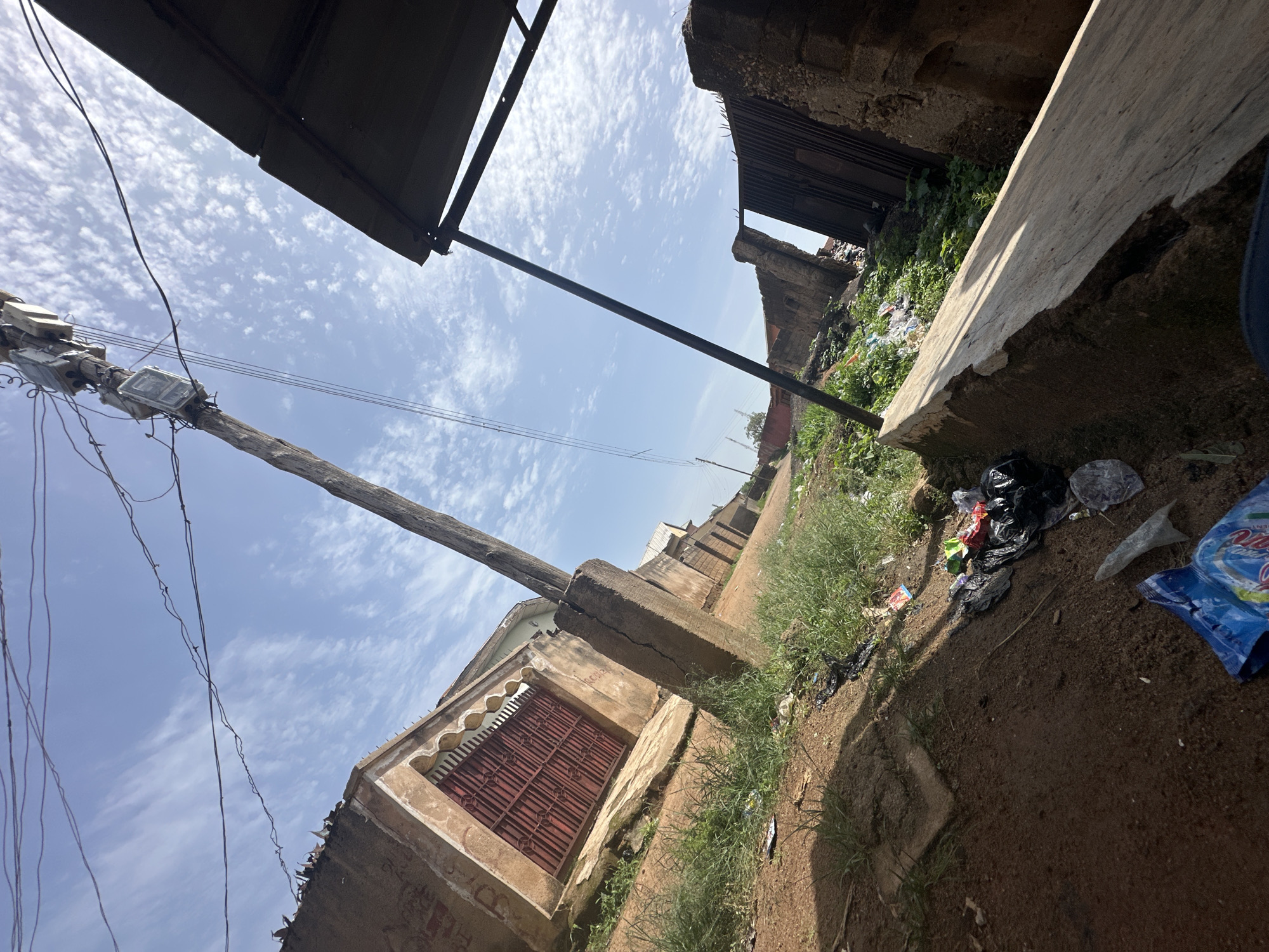 The image depicts an outdoor scene in an urban environment, likely in Jos, Nigeria, characterized by a mix of dilapidated structures and accumulated litter. Dominating the foreground and stretching into the midground is a dirt pathway with sparse, overgrown grass. Scattered across this pathway and along the edges of the surrounding structures are various forms of refuse, including plastic bags, wrappers, and indeterminate debris.
To the left, a building with rough-hewn, tan-colored walls is visible, featuring a large, red metal gate with intricate decorative elements. A concrete pillar, cracked and weathered, stands prominently in the foreground, supporting overhead electrical wires that traverse the sky. Further into the midground, another building with similar construction is seen, partially obscured by foliage.
On the right side of the image, a dark, corrugated metal roof is supported by a metal pole, casting a shadow. Adjacent to this is a structure with crumbling brick walls and a rusty, vertical metal gate, suggesting a state of disrepair. In the background, more buildings and vegetation are visible, with a clear blue sky punctuated by scattered white clouds. The overall impression is one of an unkempt urban area, possibly a residential or commercial zone, showing signs of neglect and inadequate waste management. There are no people visible in the frame.