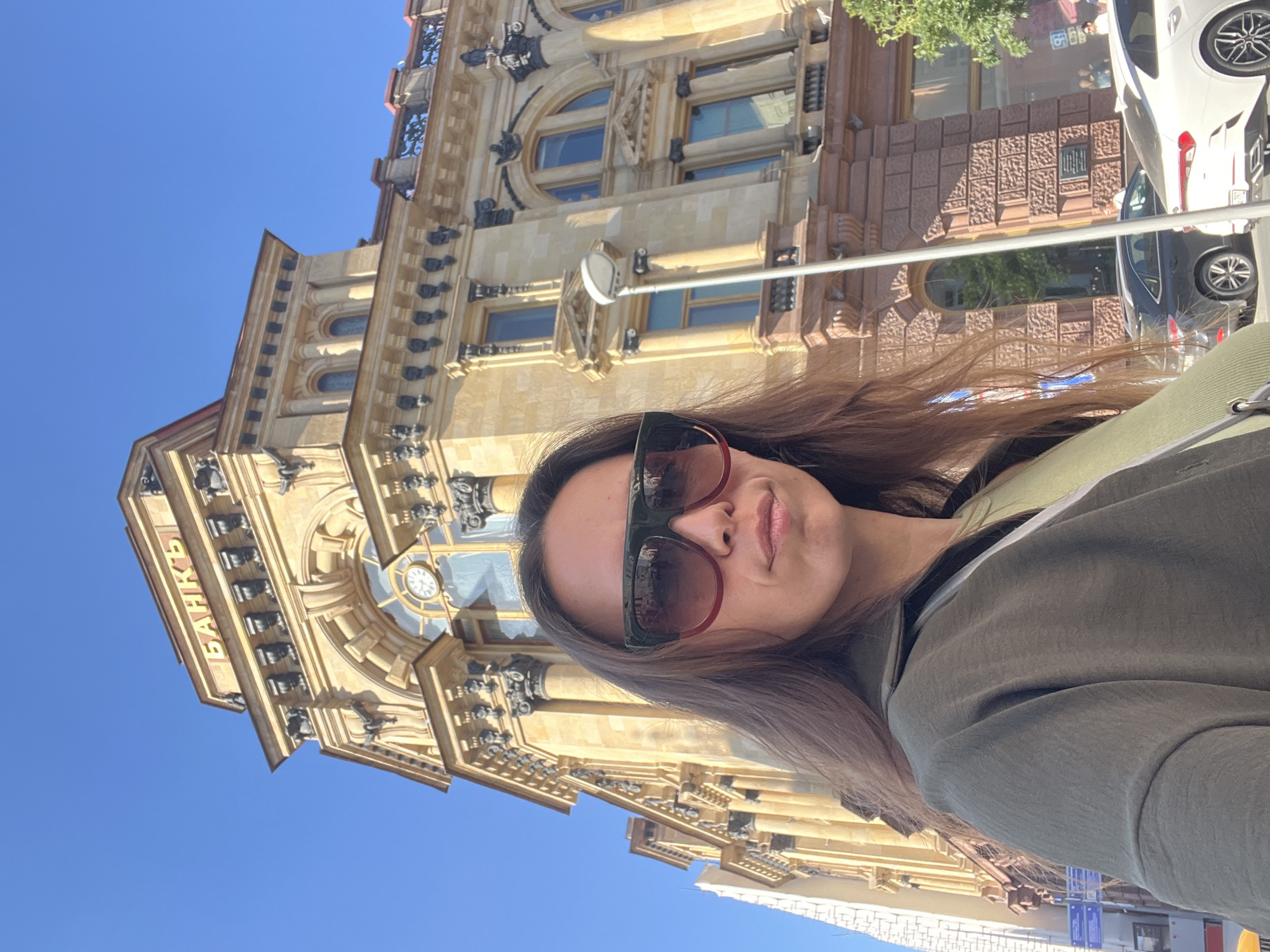 A woman wearing oversized sunglasses and a green jacket takes a selfie in front of a grand, old bank building in Moscow, Russia. The building, made of light-colored stone with ornate architectural details, has a sign in Cyrillic that reads "БАНКЪ" (BANK). The sky is a clear, bright blue. The woman is positioned in the foreground, with the building occupying most of the background. Several cars are visible on the street below.