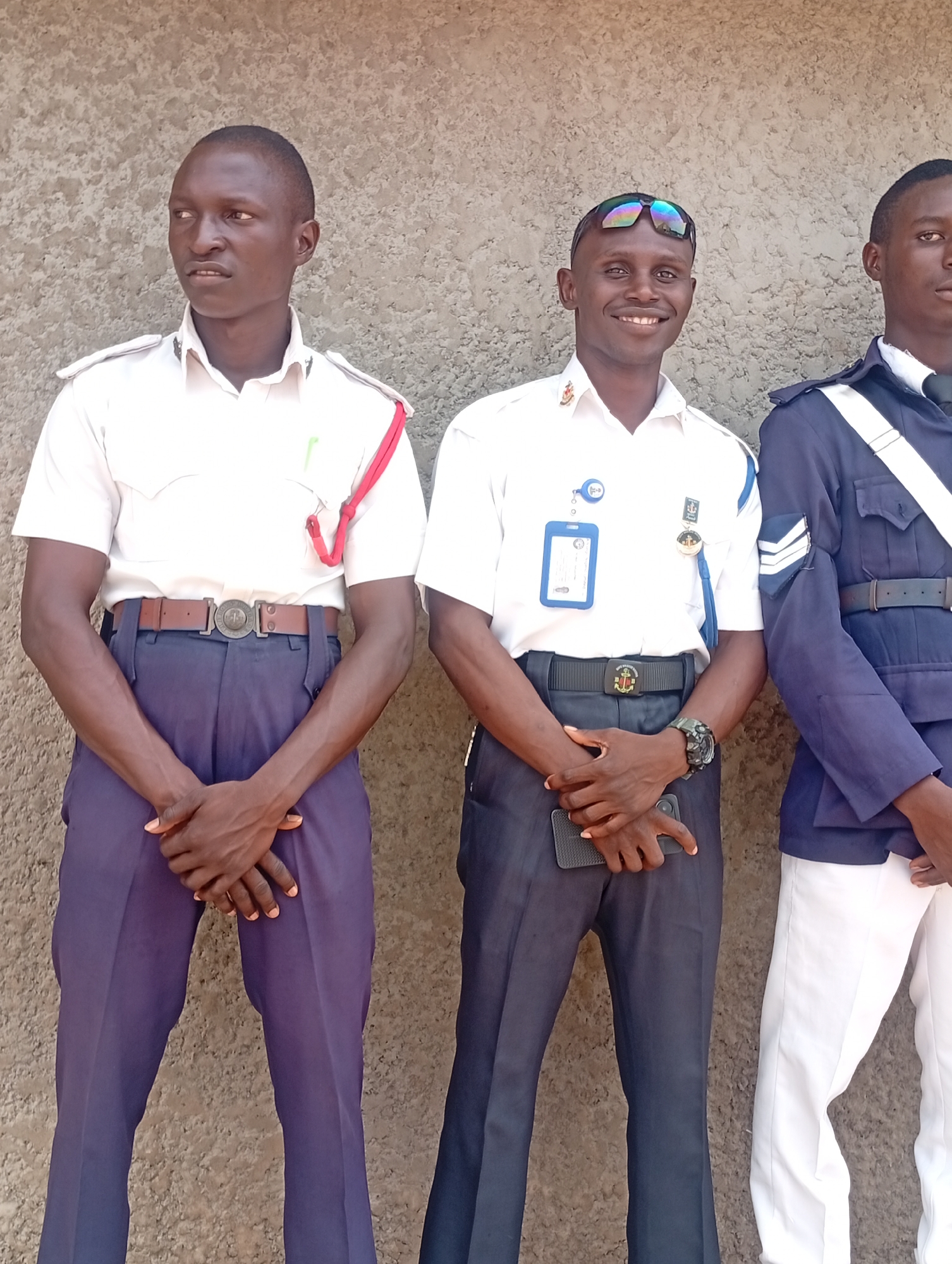 Three men in uniforms stand in front of a concrete wall in an outdoor setting. The man on the left wears a white short-sleeved shirt, dark trousers, and a brown belt with a circular buckle; a red cord hangs from his shoulder. The man in the middle wears a similar white shirt, dark trousers, a black belt with a circular buckle, and sunglasses atop his head. He also has a name tag and a watch on his left wrist, holding a cell phone in his left hand. The man on the right wears a navy blue jacket, white trousers, and a black belt. The location is in the city of Unknown, Unknown.