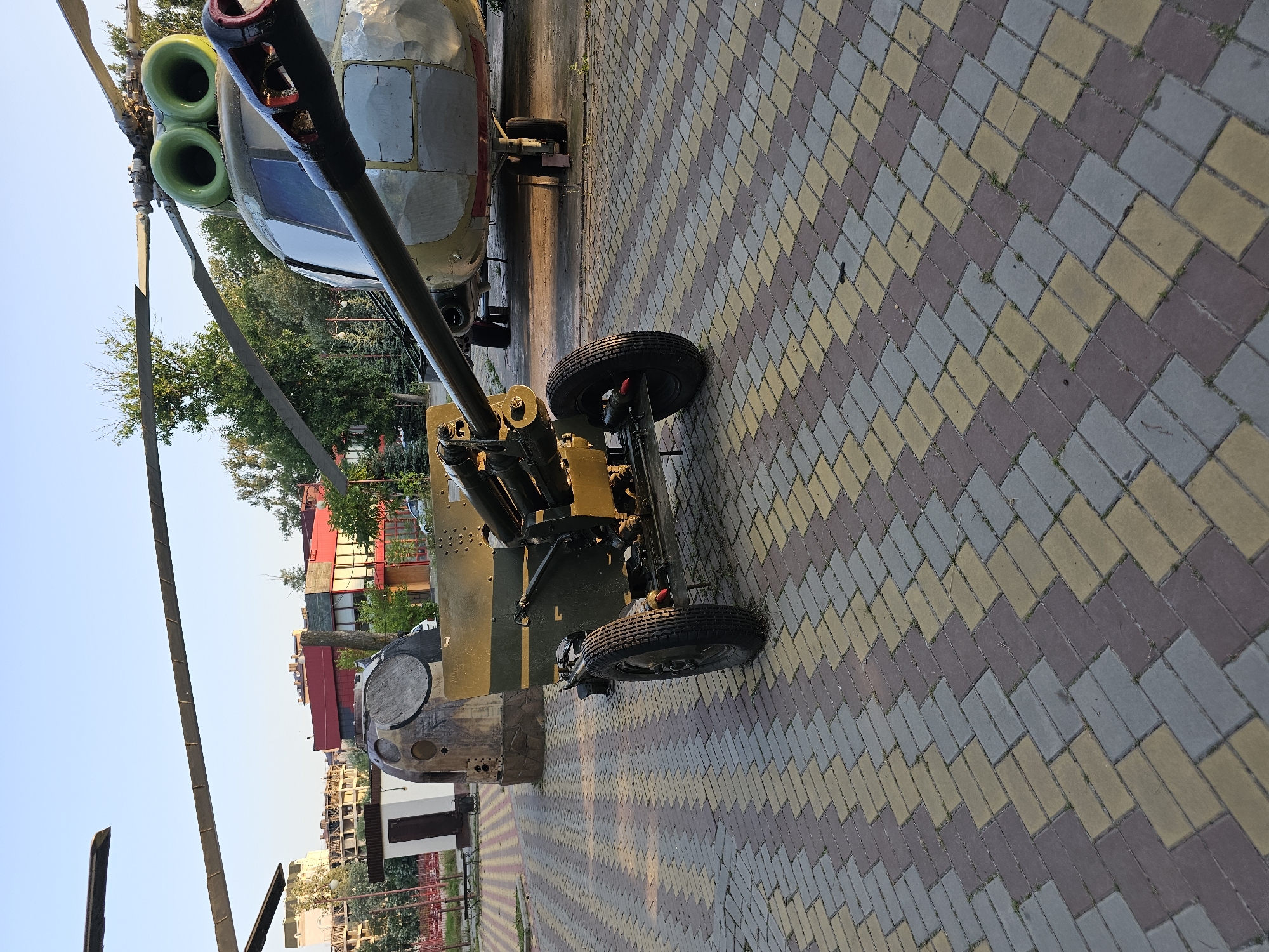 The image depicts an outdoor exhibit, likely in a museum or public space, featuring military equipment. In the foreground, a towed artillery piece, painted in camouflage green and yellow, is positioned on a patterned paver surface. The artillery piece has two wheels and a long barrel that extends towards the right side of the frame. Behind the artillery, a large helicopter, predominantly light gray with green engine intakes, is visible. The helicopter's rotors are extended. In the background, there are buildings, trees, and a large, dome-shaped structure that appears to be part of the exhibit. The scene is bathed in the warm light of what appears to be late afternoon or early morning, casting shadows on the ground. The overall environment suggests a clear day with a bright sky. The location is identified as Cherkessk, Russia. No people are visible in the image.