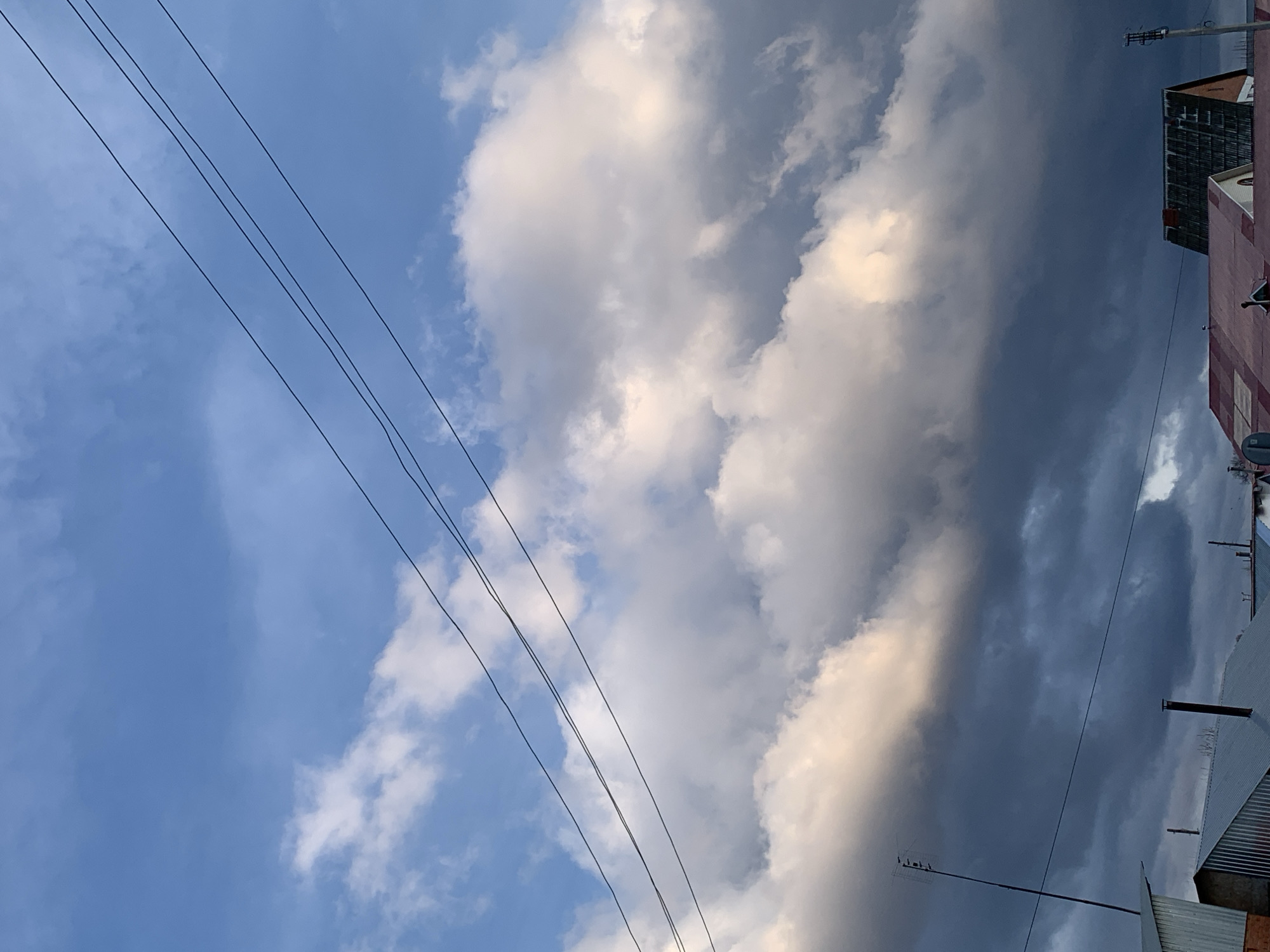 The image captures a dynamic outdoor scene in Фоки, Russia, featuring a sky filled with dramatic clouds and prominent utility lines above urban structures. The upper and central parts of the frame are dominated by the sky, which displays a striking contrast between clear blue patches on the left and a vast expanse of clouds across the middle and right. These clouds range from bright, sunlit white and light gray to dense, dark gray formations, suggesting either late afternoon lighting or impending weather changes.
Several dark utility wires, likely power lines, stretch diagonally across the upper-left and central portions of the sky. On the right side and bottom-right, sections of urban buildings are visible. One structure features a distinct reddish-pink facade, while another appears with a darker, possibly corrugated metal or weathered finish. Rooflines, windows, and an antenna on a rooftop are among the discernible architectural details. The scene is static, devoid of people or specific activity, and no text is visible. It presents a common urban vista highlighting the interplay between natural skyscape and man-made infrastructure.