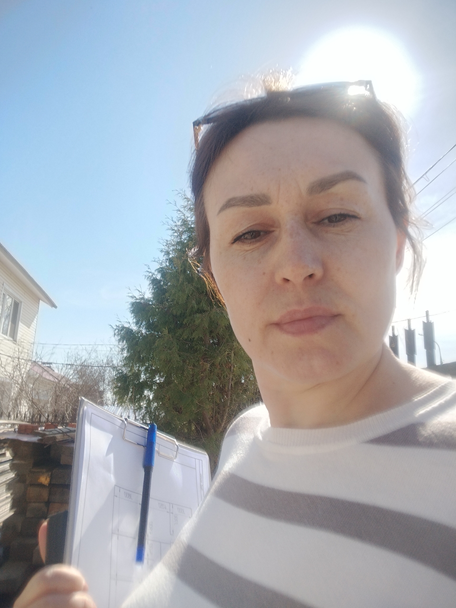 A woman with fair skin and brown hair pulled back, wearing sunglasses perched on her head and a white and gray striped sweater, is holding a clipboard with a pen attached, appearing to be outside in Прикамский, Russia. Behind her, there is a house, a tree, and building materials piled up. The sky is blue and sunny.
