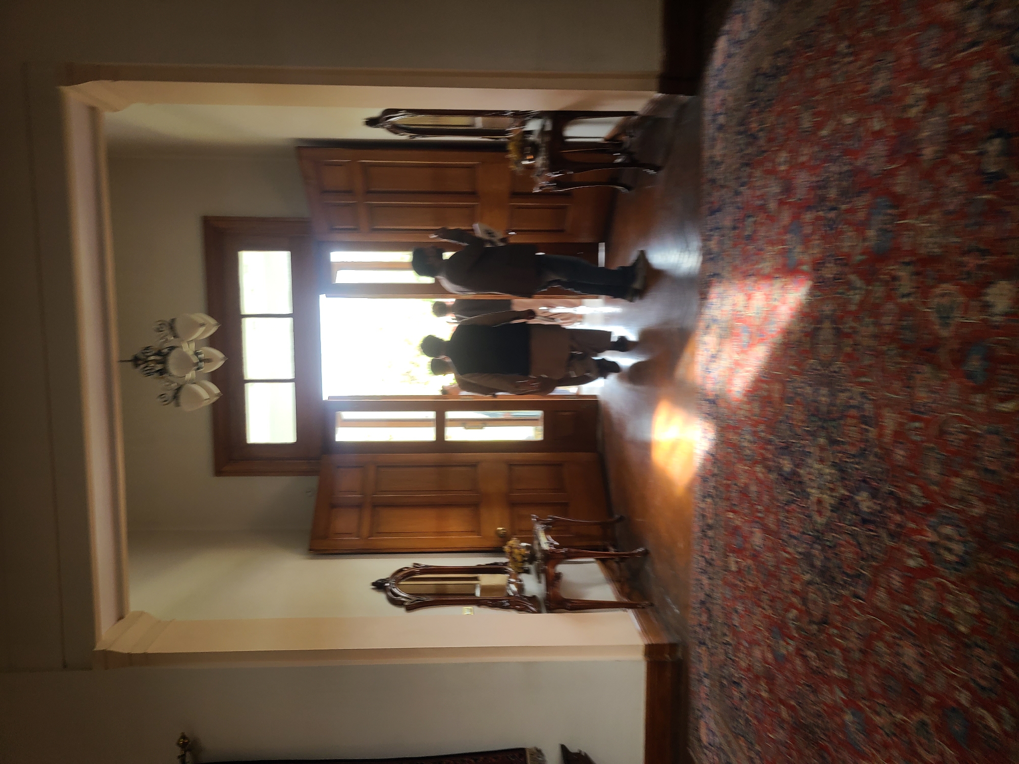 The image captures an interior view of a building, likely a grand residence or public space, in Quetta City Tehsil, Pakistan. The perspective is from inside, looking towards a bright exterior through an arched doorway.
In the center, several people are silhouetted against the bright natural light flooding through large, paneled wooden double doors, which feature glass panels at the top. They appear to be standing in the doorway, possibly entering or exiting. Their faces and specific details are obscured by the strong backlighting.
To the right of the doors, against a light-colored wall, an ornate wooden console table with cabriole legs and a matching decorative mirror above it adds to the room's traditional aesthetic. On the left side of the room, a multi-branched chandelier hangs from the ceiling.
The floor is made of polished wood, partially covered by a large, intricately patterned rug predominantly in red and blue hues. Bright sunlight streams through the doorway, creating strong highlights and shadows across the wooden floor and the rug. The overall impression is one of an elegantly furnished space, illuminated by clear daytime light. There is no visible text.