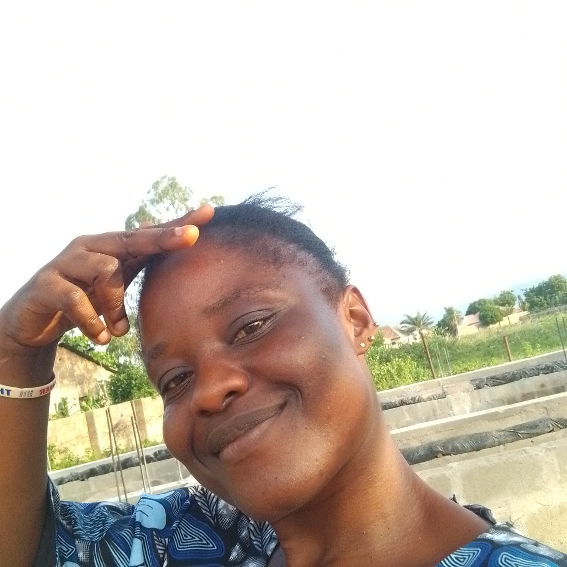 A woman is taking a selfie outdoors. She has dark skin and short, dark hair. Her left hand is raised and touching her forehead. She is wearing a blue and white patterned shirt and a white bracelet with black writing. She has small gold earrings in her left ear. The background shows a somewhat overgrown, rural scene with buildings and vegetation. There are also some concrete structures that appear to be part of a construction or agricultural setup, with black coverings. The lighting suggests it is daytime, likely late afternoon, given the warm light and shadows. The overall emotion conveyed is one of happiness and warmth, as she is smiling broadly. The bracelet on her wrist has visible text, which appears to be "OWER" and some other illegible words. The location is Jalingo, Nigeria.