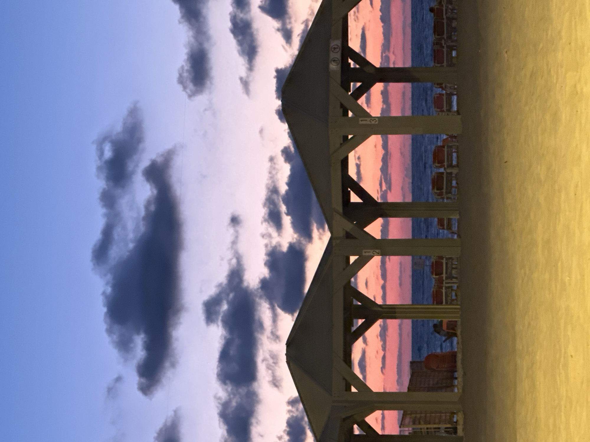 The image shows a beach scene at sunset in Tel Aviv, Israel, viewed through the wooden structures of beach canopies. The sky is a mix of blues, pinks, and grays with scattered clouds. The sea is visible as a dark horizontal line through the canopies. Beach chairs are lined up behind the canopies. The sand is visible in the foreground. A person is sitting in the shade of one of the canopies.