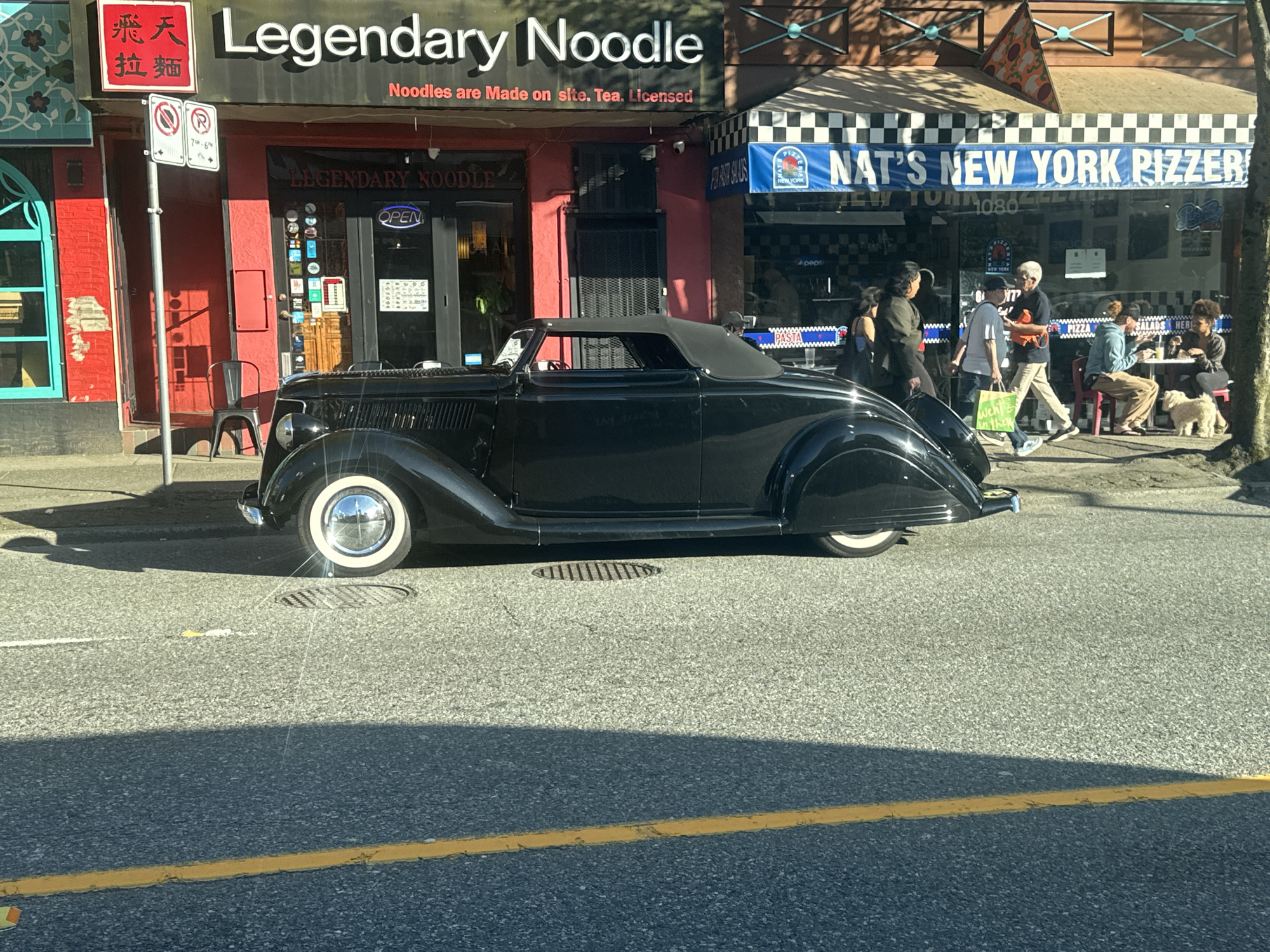 The image captures a scene on a street in Vancouver, Canada, featuring a black vintage convertible parked in front of two businesses. On the left is a restaurant called "Legendary Noodle," with Chinese characters visible on a sign above the door. Adjacent to it is "Nat's New York Pizzeria," with a blue and white awning. Several people are walking by or seated outside the pizzeria. The street is paved, with a yellow line marking lanes and a manhole cover near the car. There is a parking sign. The lighting suggests it's daytime, with shadows cast on the street.