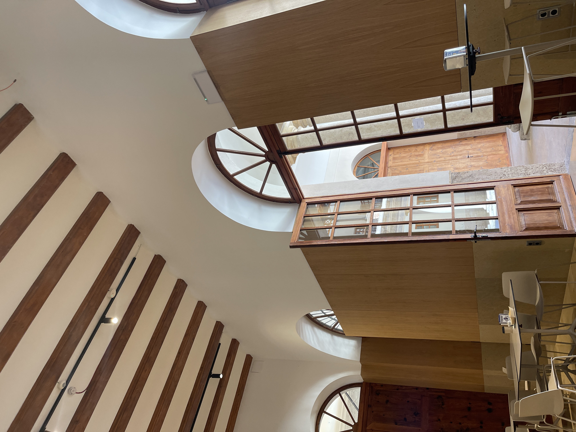 The image shows an interior view of a building in València, Spain. The camera is pointed upwards towards a ceiling with wooden beams and arched windows. The architecture is a mix of old and new, with the wooden beams suggesting an older structure and the arched windows providing natural light. A modern wooden structure is visible in the center of the image, extending horizontally and obscuring part of the ceiling and windows. This structure has a smooth, light-colored surface and appears to be part of a more recent renovation. White walls are visible, contrasting with the warm tones of the wood. Artificial lighting fixtures are attached to some of the wooden beams. The scene is well-lit and appears to be taken during the daytime. The architecture and design suggest a building with historical significance that has been renovated for contemporary use.