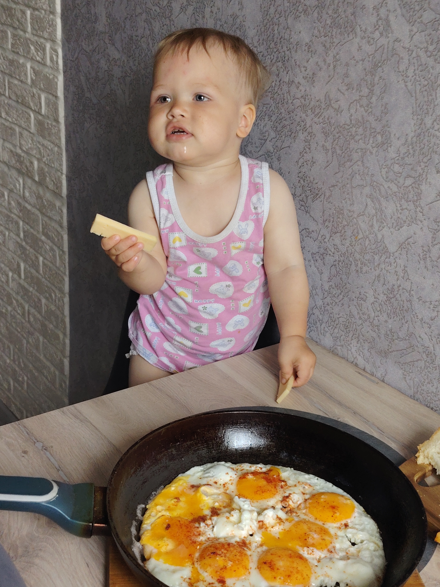 A young child, possibly a toddler, stands at a table in what appears to be a kitchen or dining area. The child is wearing a pink tank top with white patterns. The child has a piece of cheese in one hand and a smaller piece of cheese on the table near a frying pan containing several fried eggs, seasoned with paprika. The location is likely in Chaikovsky, Russia, based on user request. The room has a brick-patterned wall on one side and a textured wall on another side. The table has a light wood-grain finish. The child appears to be observing the food.