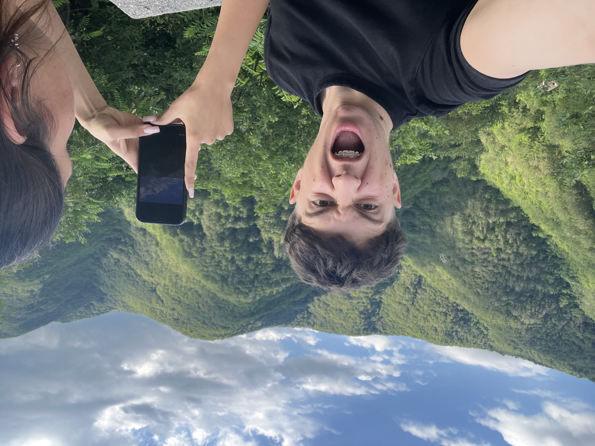 In an outdoor scene set against a backdrop of lush, green mountains and a partially cloudy blue sky, a young person with dark, curly hair and braces is making a comical face directly at the camera. They are wearing a black t-shirt. To the right, another person's hands are visible, holding a black smartphone and taking a picture of the mountainous landscape. The location appears to be in or near Pechinska, Bulgaria. The overall atmosphere is informal and adventurous.