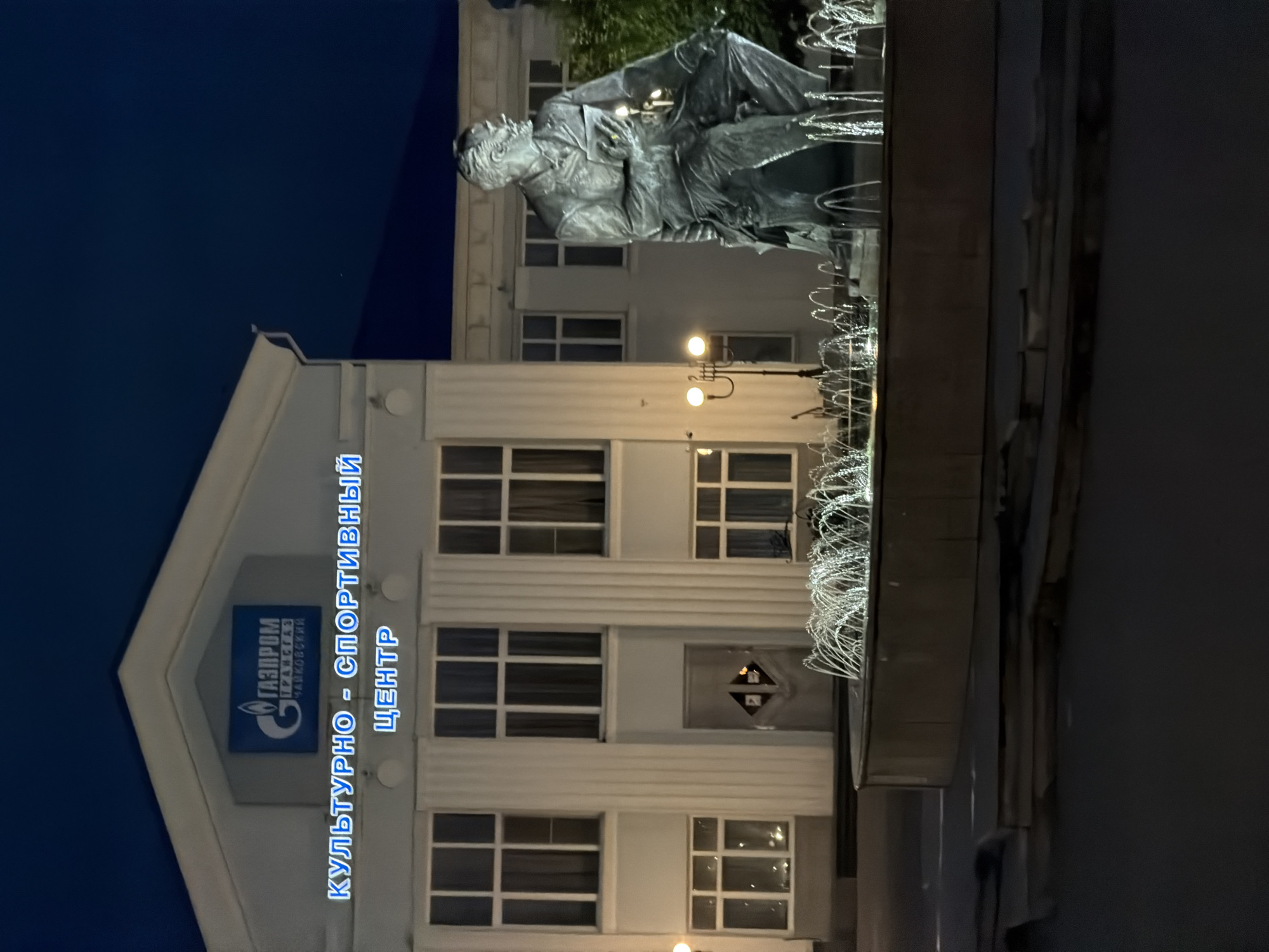 The image depicts the "Gazprom Transgaz Chaikovsky" Cultural and Sports Center in Chaikovsky, Russia, at night. A statue of a seated man is prominent in front of the building, with a fountain featuring illuminated water jets in the foreground. The building itself is well-lit, highlighting its architectural features, including large windows and columns. The sky is dark, indicating nighttime. The overall scene is illuminated by artificial lights from the building, the fountain, and a streetlamp visible to the left of the statue.