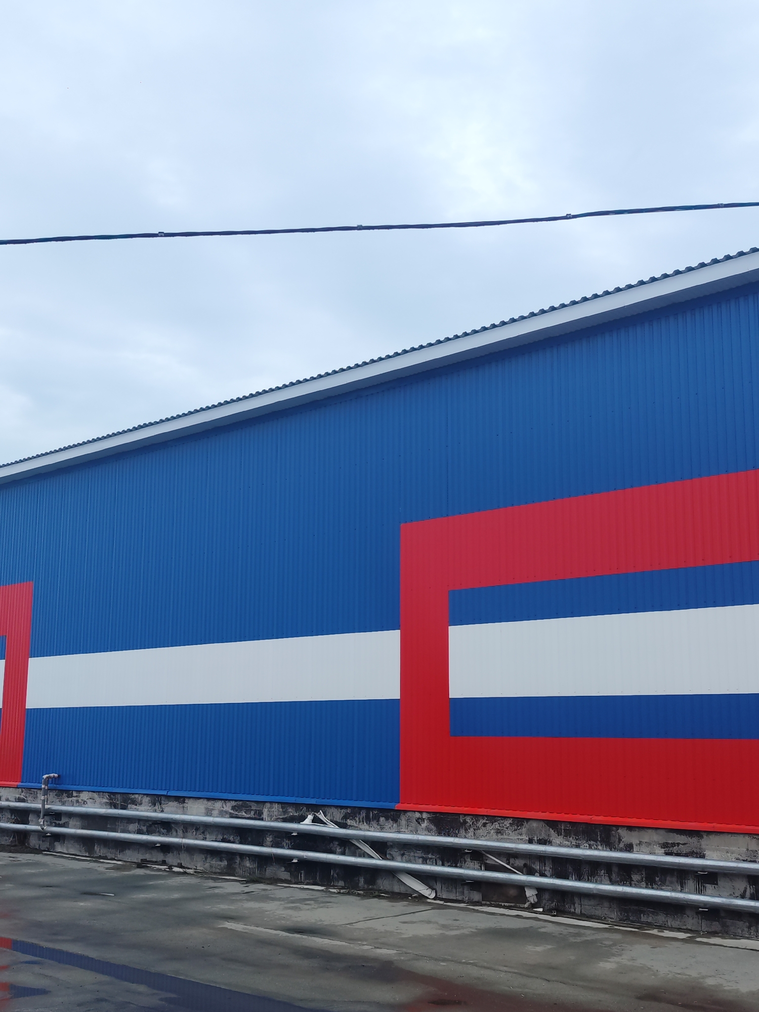 The image shows the exterior of a building in Chaikovsky, Russia. The building's facade is made of corrugated metal panels painted in a pattern of horizontal stripes of blue, white, and red. A red, geometric shape partially frames the right side of the white stripe. An electrical line stretches across the upper part of the frame against a gray sky. A metal pipe runs along the base of the building, next to a concrete surface.