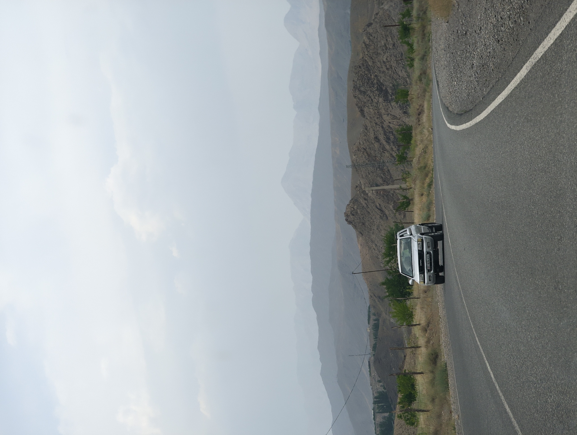 A silver Opel SUV is driving on a paved road in a mountainous region. The car is white with a license plate that reads "EA 634 ACG". The road curves to the right, and there are dry grass, scattered trees, and fences along the side. In the background, there are several layers of hazy, muted mountains. The sky is overcast and light. The image appears to be taken from another vehicle on the road. The location is described as being in the city of Samarkandyk, Kyrgyzstan, which is known for its mountainous terrain and remote roads.
