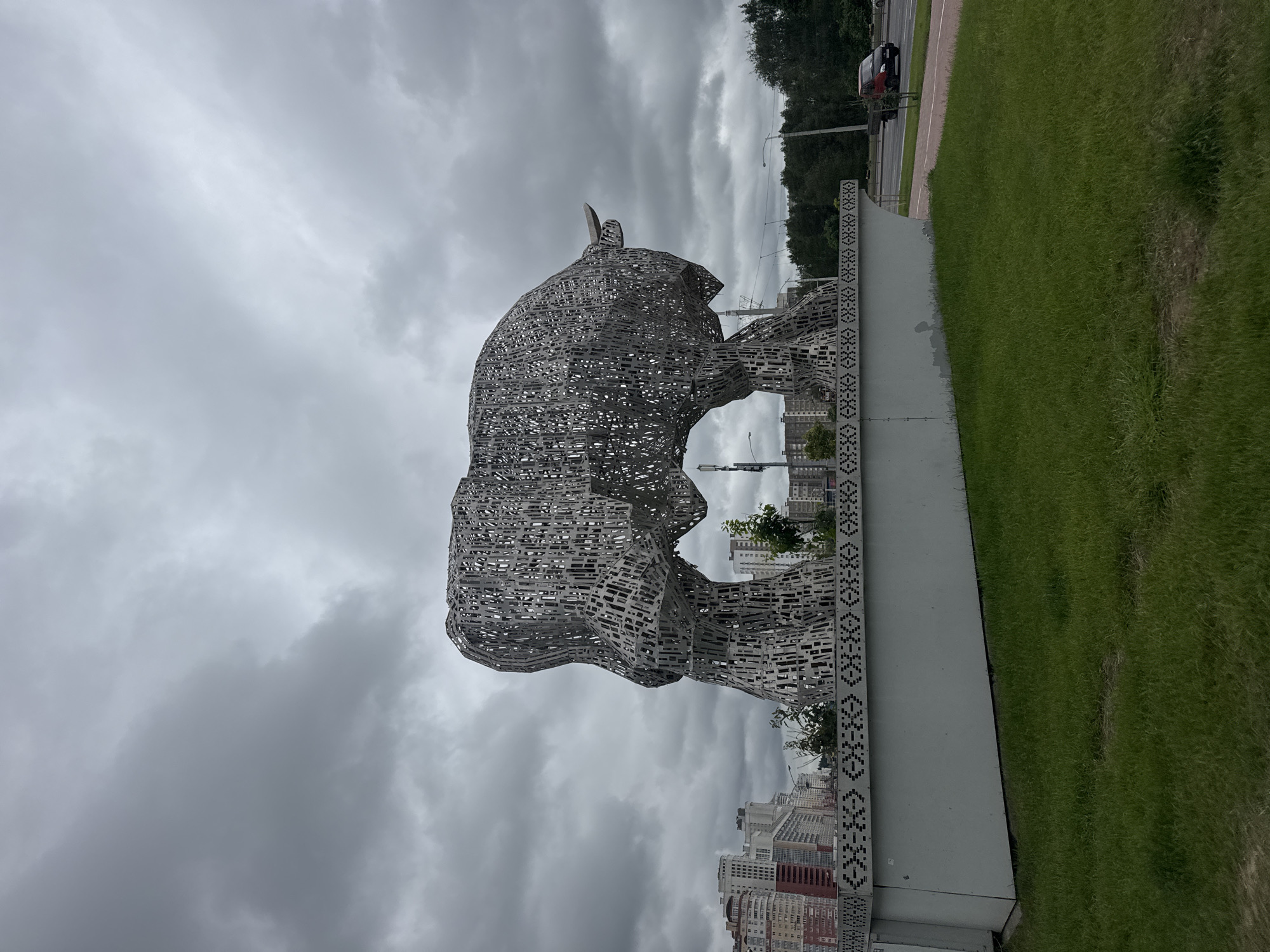 An outdoor, low-angle, full shot captures a large, abstract metal sculpture of a bull under a cloudy, overcast sky. The sculpture is constructed from numerous metallic rectangular pieces, forming an open, intricate design. It stands on a white, rectangular base that is adorned with a geometric pattern. The bull sculpture is positioned in the foreground, with a city skyline featuring tall, multi-story buildings visible in the background. Green grass covers the ground in front of the sculpture's base. To the right of the sculpture, a road with passing traffic, including a red car, can be seen. The environment suggests an urban park or public space in Minsk, Belarus.