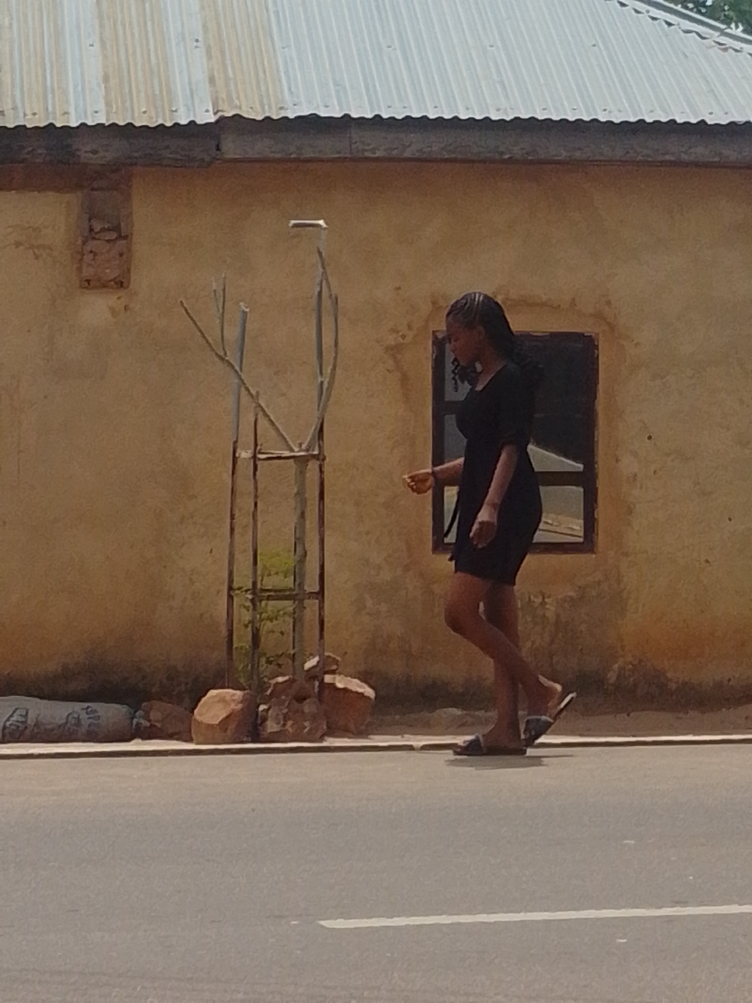A young woman in a black dress and sandals walks along a paved road in front of a tan building with a corrugated metal roof. A small tree in a metal cage stands to the left of the woman, with several rocks at its base. A large, dark, cylindrical object is also present to the far left of the frame, partially obscured. The building has a single, dark-framed window. The road has a white line marking its edge.