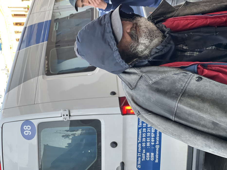 The image captures an outdoor urban scene in Tanger, Morocco, near the ZI Voie 21 on the Tetouan route, as indicated by signage on a vehicle. The primary subject is a male figure, positioned in the foreground, slightly right of center, seen in profile facing left. He has a prominent, unkempt grey and white beard and is dressed in multiple layers: a dark blue hooded jacket with the hood up, a grey baseball cap worn under the hood, a weathered grey overcoat, and a red padded vest over a dark plaid shirt. His gaze is directed forward and slightly downward. Partially visible to the right of the primary subject is a second male, with only the right side of his face and shoulder discernible. This individual has short dark hair, wears a dark cap, and holds his right hand near his mouth. The background features a white utility van with horizontal blue and grey stripes, tinted windows, and a blue oval decal marked "98" on its rear door. The van's side also displays a blue sticker with company details for "IBRATRANS," including "ZI Voie 21 route Tetouan, Tang" and contact numbers. Distant, blurred urban architecture is visible above the van. The scene is illuminated by daylight. No direct interaction between the two visible individuals is depicted.