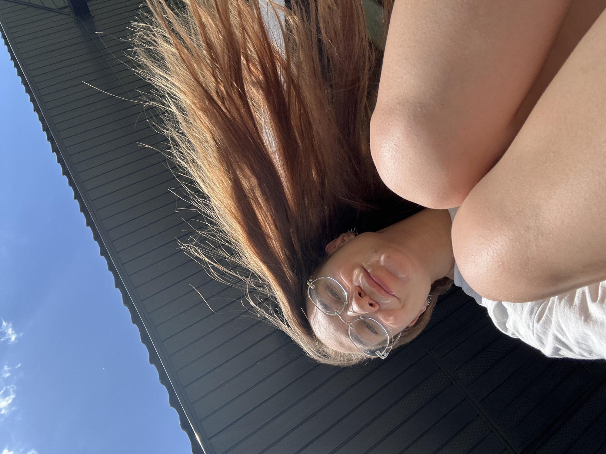 A young woman with long, light-brown hair is looking down at the camera, upside down. She is wearing round, wire-rimmed glasses and a white shirt. Her arms are crossed in front of her. The background consists of a gray metal roof and a blue sky with a few clouds. The image does not show any recognizable landmarks or specific details that would indicate it was taken in Kharnavy, Russia.