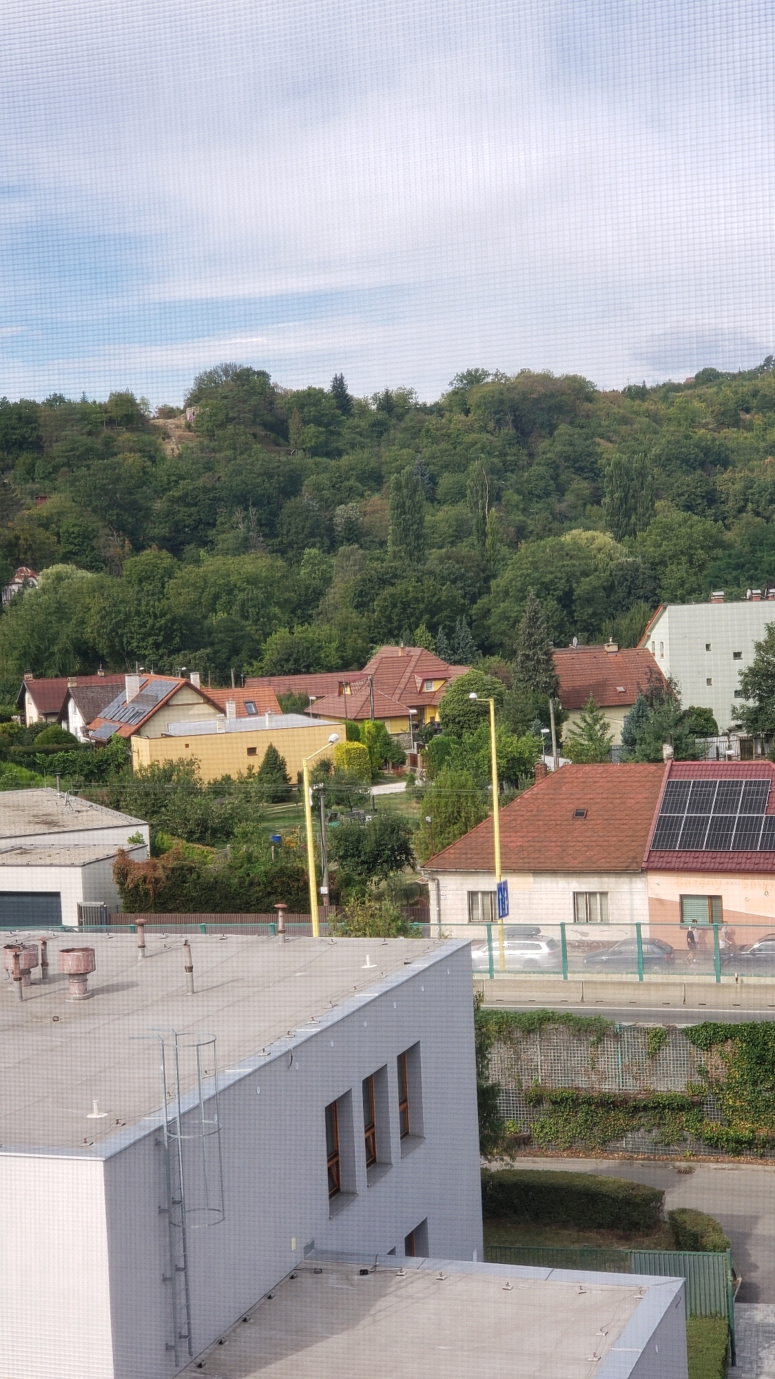 This is an outdoor, daytime, medium-shot photograph taken from a slightly elevated perspective, looking towards a suburban residential area backed by a wooded hill. In the foreground, the roof of a modern, light-colored industrial building is visible, featuring multiple ventilation pipes and a metal ladder attached to its side. To the right and behind this building, a multi-story apartment complex with white siding and multiple windows can be seen. Further back, a street with a few cars and pedestrians is bordered by a retaining wall covered in green ivy. Beyond the street, a cluster of houses with red-tiled roofs and yellow and white exteriors are nestled amongst trees. A tall, yellow lamppost is prominent in the mid-ground. The background is dominated by a densely forested hillside under a partially cloudy, light blue sky. The overall scene depicts a quiet, suburban environment with a mix of residential and industrial buildings set against a natural landscape. The image does not contain any specific landmarks or visual cues that definitively place it in Prešov, Slovakia, but the architectural style of the houses and the natural setting are consistent with areas in and around the city.